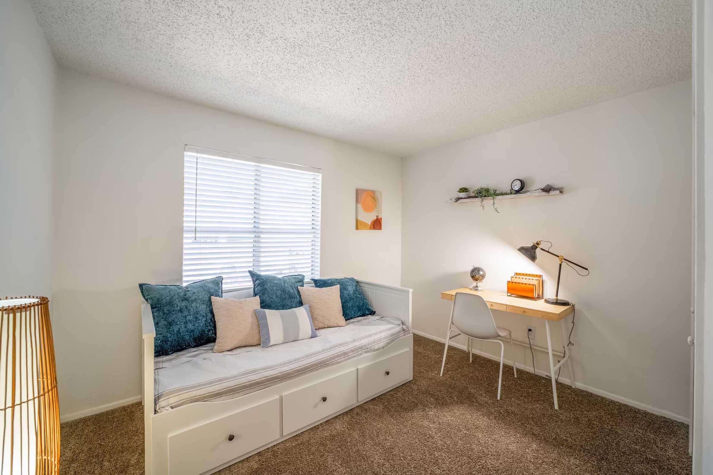 Cozy room featuring a daybed with blue and beige cushions, a desk with a lamp, and a decorative wall shelf. Natural light streams in through a window with blinds, and a simple artwork is displayed nearby. The flooring is carpeted, enhancing the room's warm and inviting atmosphere.