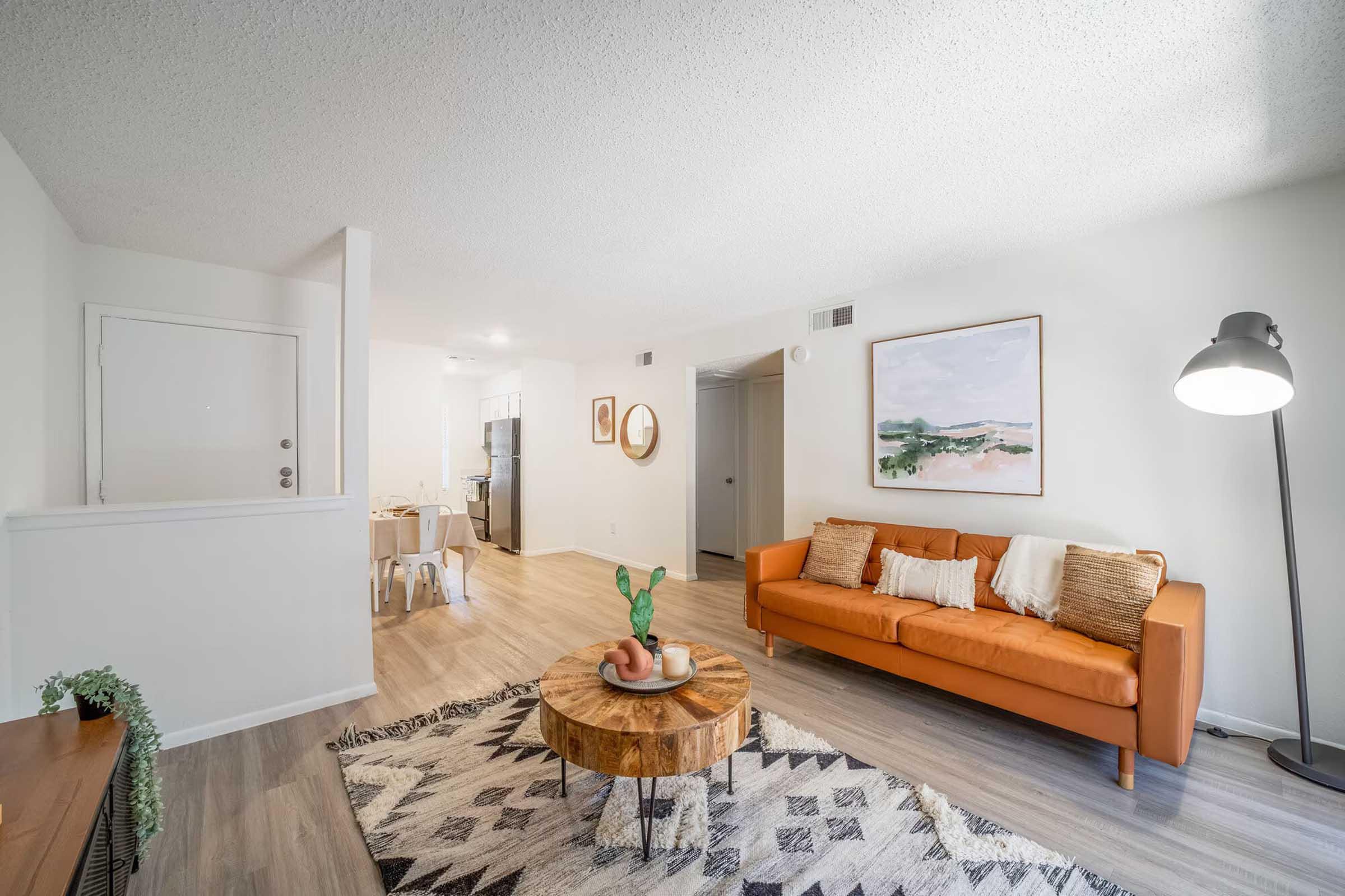 Cozy living room featuring a bright orange couch with decorative pillows, a wooden coffee table with plant decorations, and a large framed artwork on the wall. The space has light wood flooring and leads to a kitchen area visible in the background, complemented by soft lighting from a floor lamp.