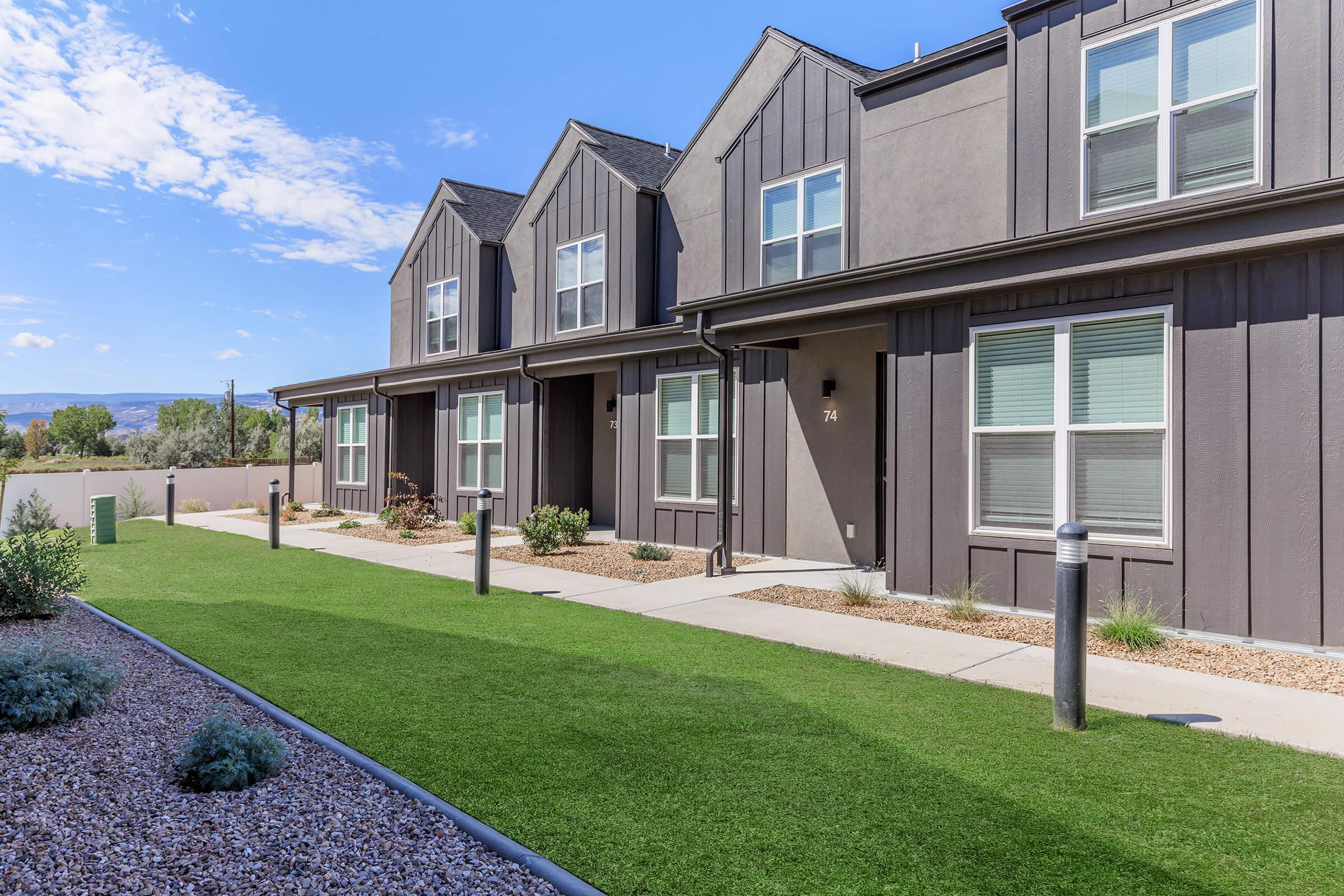 Contemporary row of multi-unit townhouses with a well-maintained lawn and pathway. The buildings have dark gray exteriors, large windows, and numbered doors. A clear blue sky with scattered clouds is visible in the background, adding to the modern aesthetic of the housing complex.