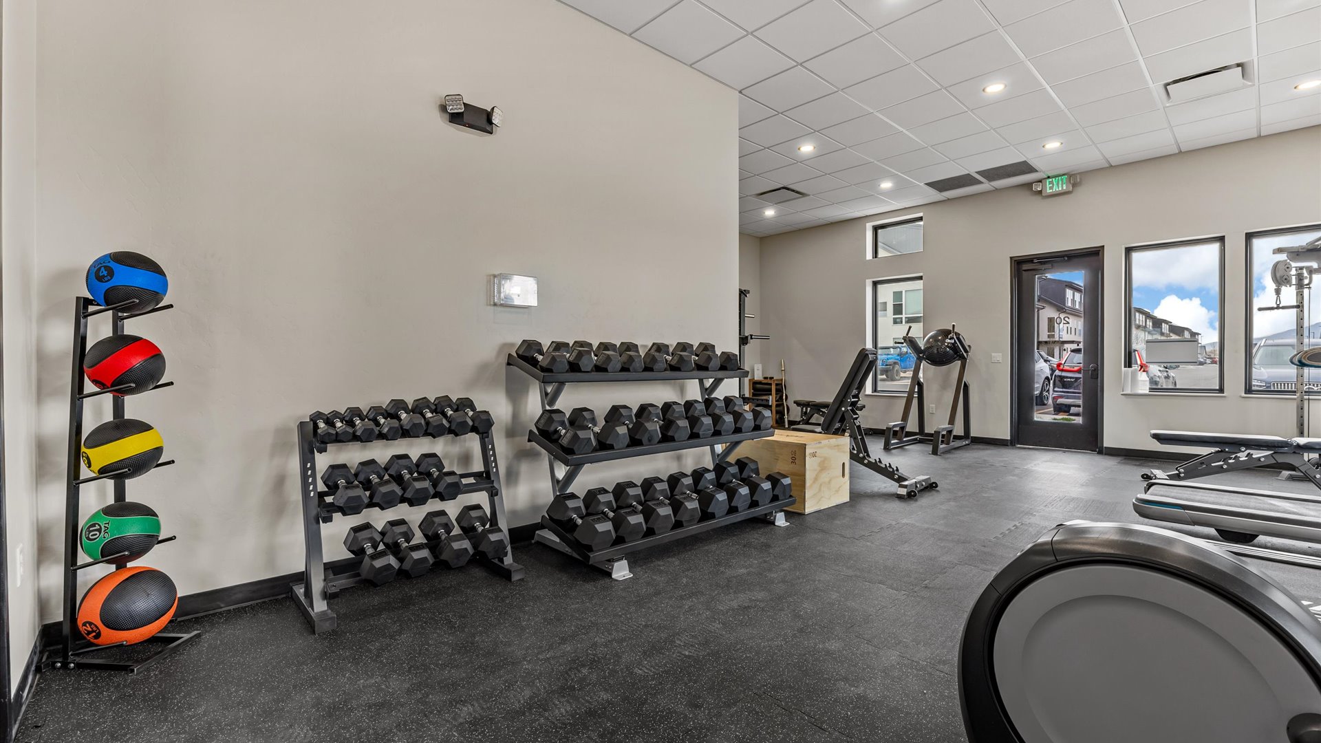 A modern gym interior featuring a variety of fitness equipment. There are dumbbells arranged on racks, a set of colorful medicine balls on a stand, a wooden box for jumps, and cardio machines near large windows allowing natural light. The floor is covered with black mats, creating an inviting workout space.