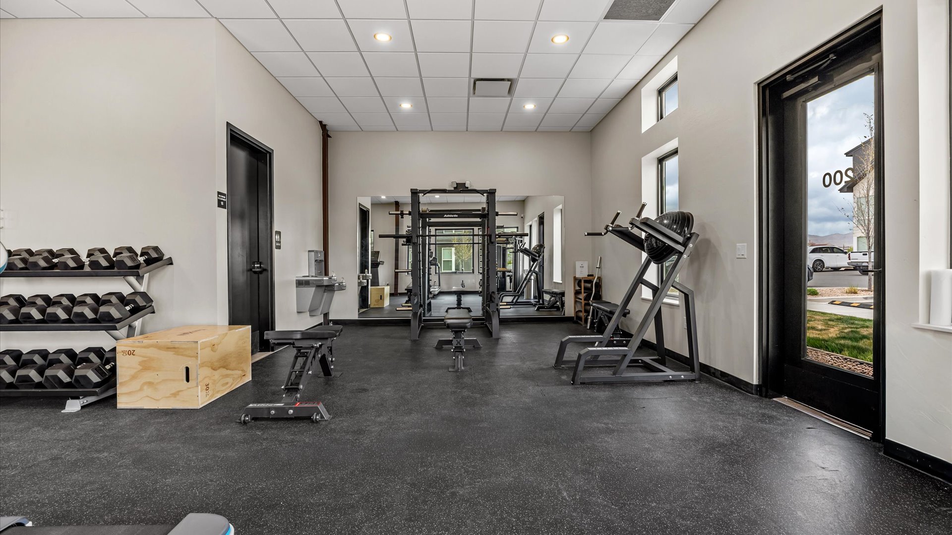 A modern gym interior featuring a variety of fitness equipment. On the left, there's a set of dumbbells on a rack, and a wooden box. Centered is a multi-functional weight machine, and on the right, there's a treadmill. The room has large windows, bright lighting, and an inviting atmosphere.
