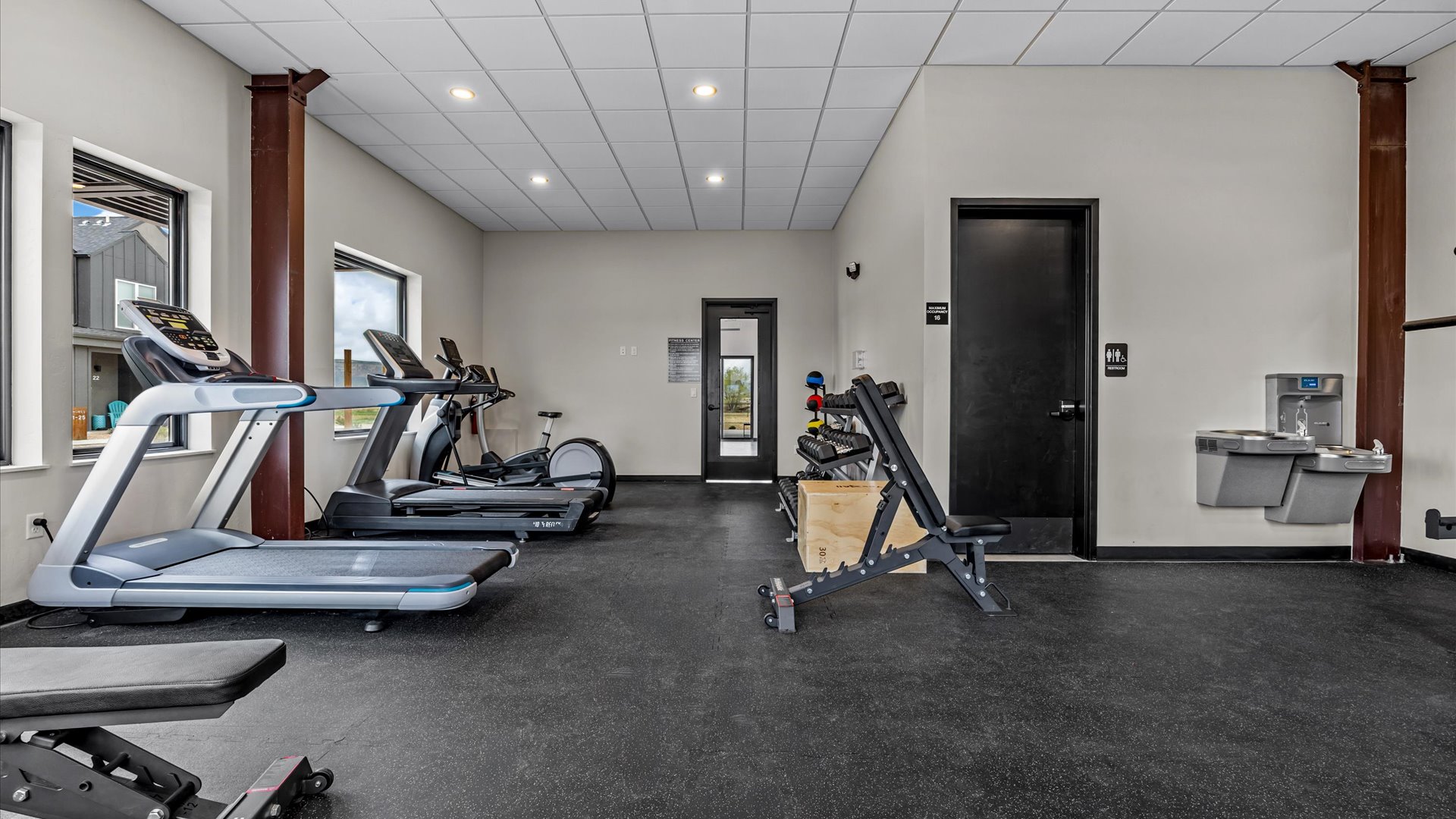 A spacious gym interior featuring multiple exercise machines, including treadmills and an exercise bike. The room has a water fountain, and a set of weights is visible on a bench. Large windows provide natural light, and the flooring is dark. The walls are neutral in color, creating a clean and modern atmosphere.