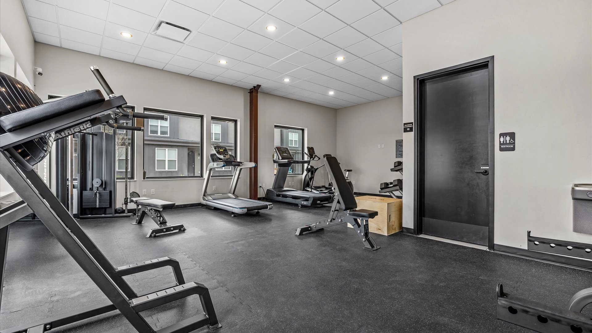 A modern gym interior featuring various workout equipment including a treadmill, weight benches, and a strength training machine. The space is well-lit with large windows, showcasing a clean, organized environment. A restroom door is visible in the background.