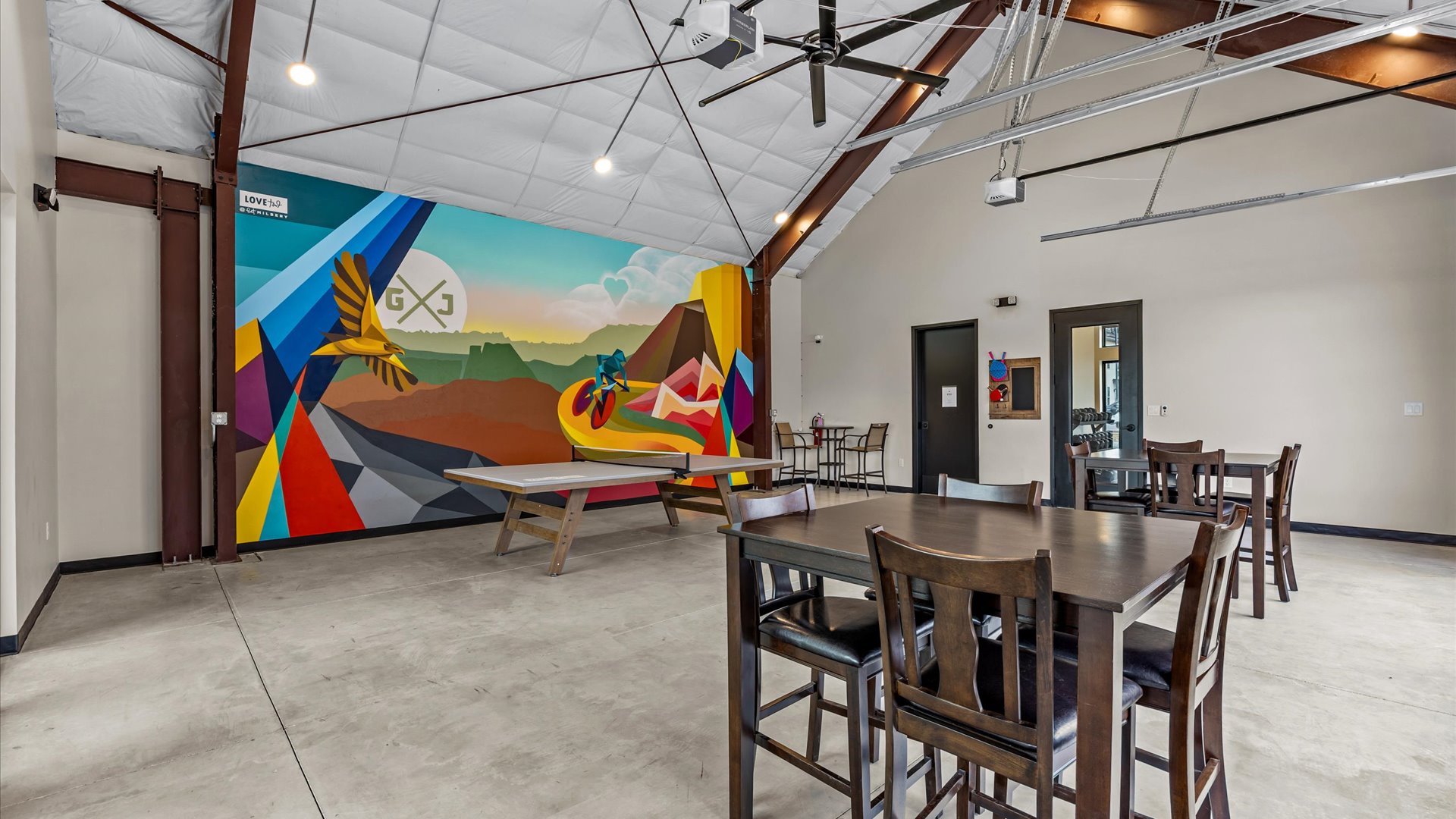 Interior of a modern recreational space featuring a colorful mural of a bird and cyclist on a wall. There is a large table tennis table in the center and several wooden dining chairs around a table. The room has high ceilings with exposed beams and a bright, open atmosphere, suitable for social gatherings.