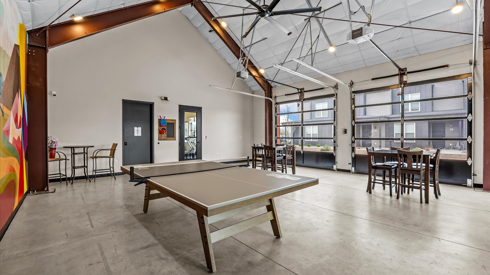 A spacious indoor recreational area featuring a ping pong table in the center, surrounded by high-top tables and chairs. Large garage doors offer natural light and a view of the outdoors. The walls are adorned with colorful artwork, creating a vibrant atmosphere.
