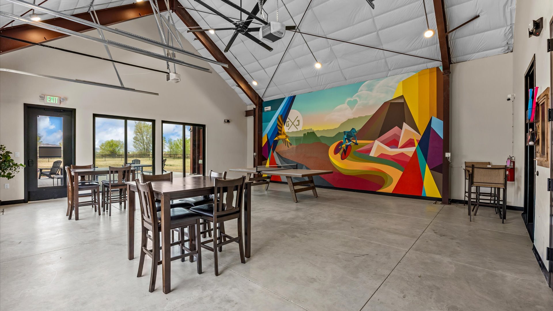 A spacious interior of a modern venue featuring high ceilings, large windows, and industrial-style decor. The room has several wooden tables and chairs arranged for seating. A vibrant mural depicting colorful geometric shapes and landscapes adorns one wall, adding artistic flair to the space.