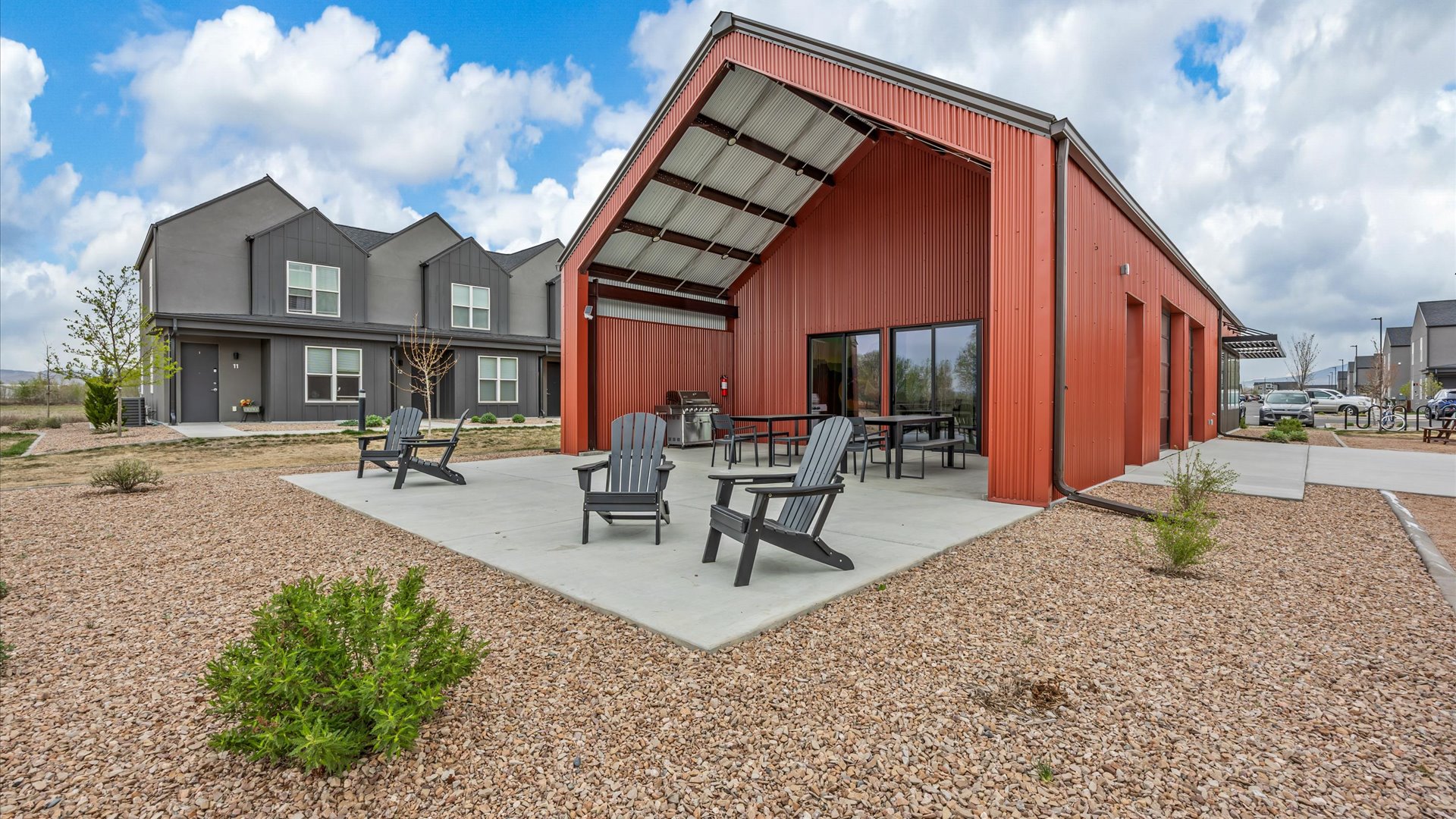 A modern red barn-style building with a large open area featuring tables and chairs. In the background, there are contemporary grey multi-family homes. The surrounding landscape includes a gravel yard with small shrubs and patches of grass, under a cloudy blue sky.