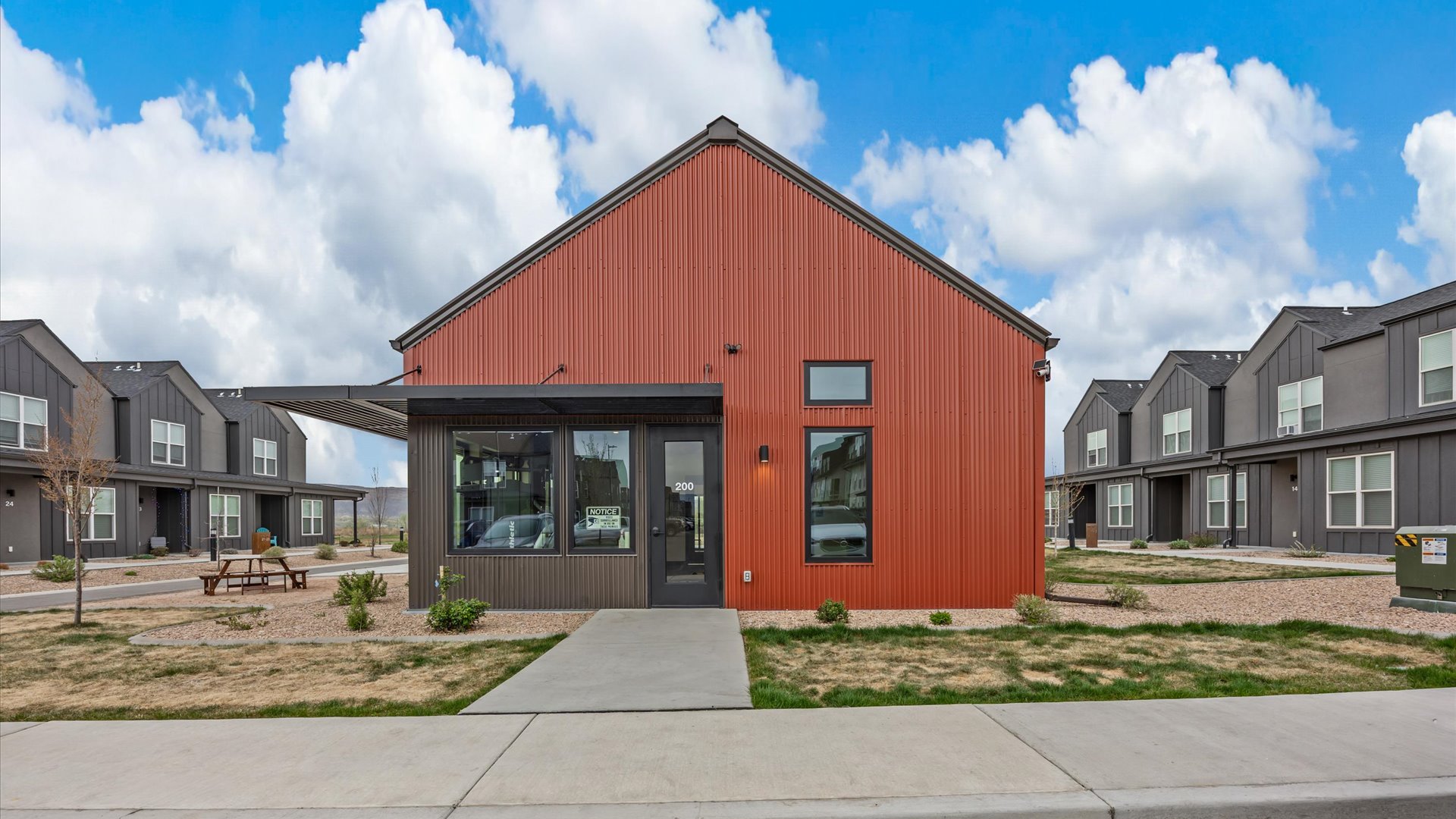 A modern single-story building with a red and gray exterior, featuring large front windows and a covered porch area. The surrounding landscape includes manicured grass and small trees. In the background, additional contemporary buildings can be seen under a partly cloudy sky.