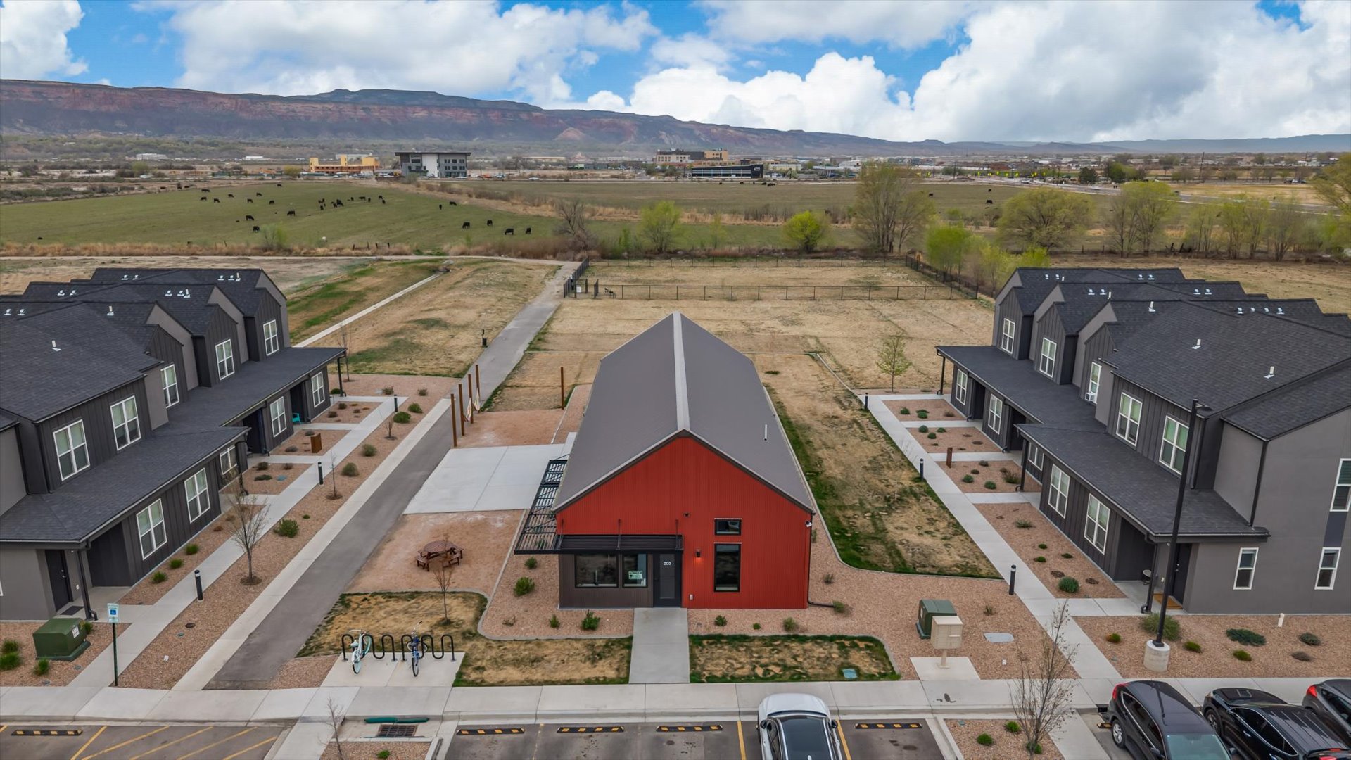 Aerial view of a modern residential complex featuring two rows of multi-unit buildings flanking a central community space with a red barn-style structure. Surrounding landscape includes open fields and distant mountains under a partly cloudy sky. Parking area visible in foreground.