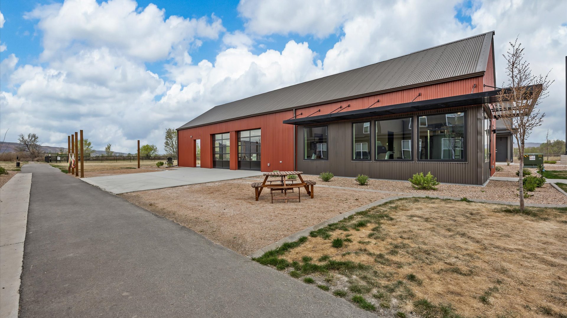 A modern red and gray building with large windows and a overhang, surrounded by a gravel area and sparse landscaping. There is a picnic table in the foreground and a paved path leading up to the building. The sky is partly cloudy, creating a bright outdoor setting.