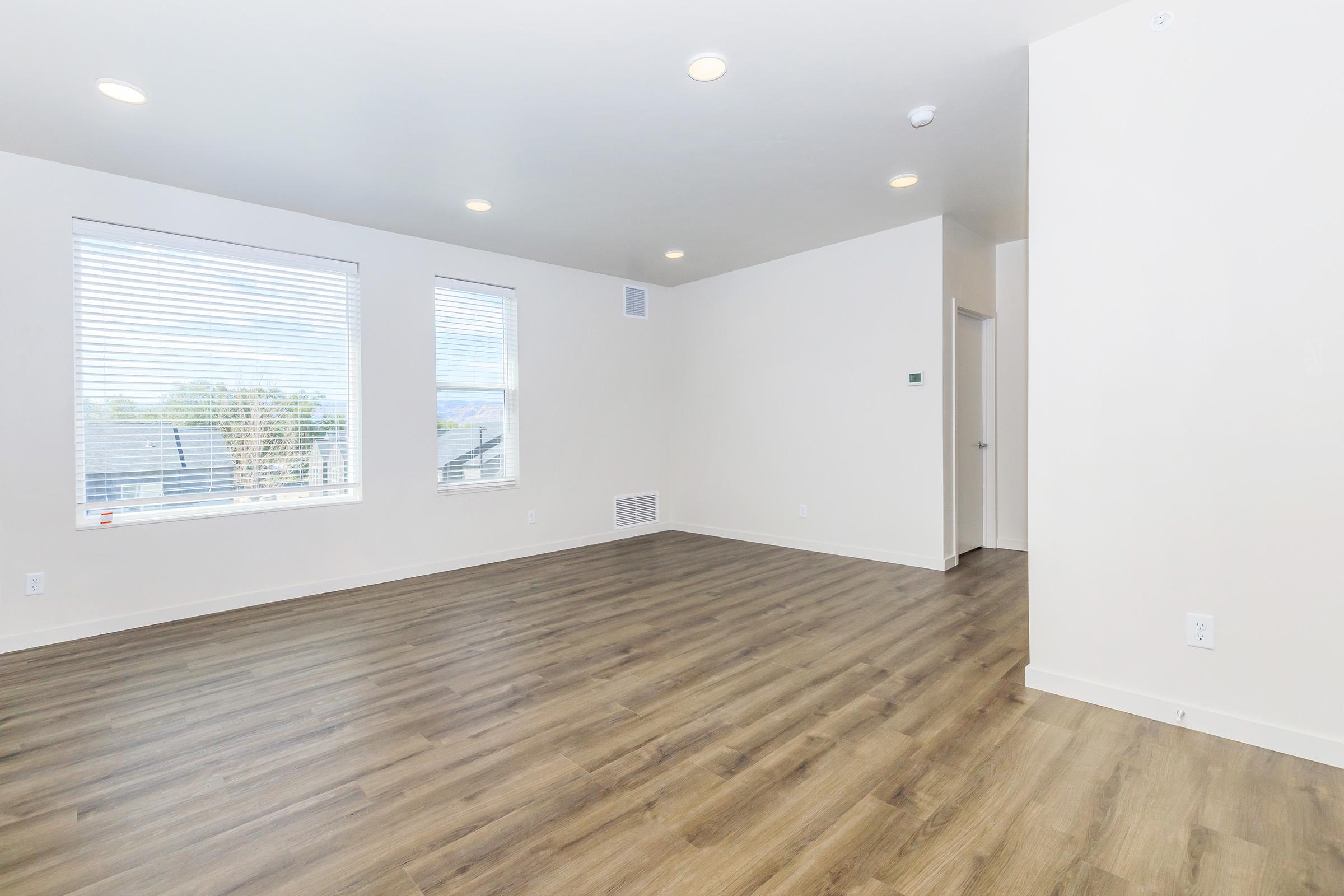 A spacious, modern living room with light-colored walls and large windows allowing natural light. The floor is made of wood laminate, and the room has recessed lighting. There is a door leading to another room and a vent on the wall, creating a clean and airy atmosphere, ideal for various furniture arrangements.