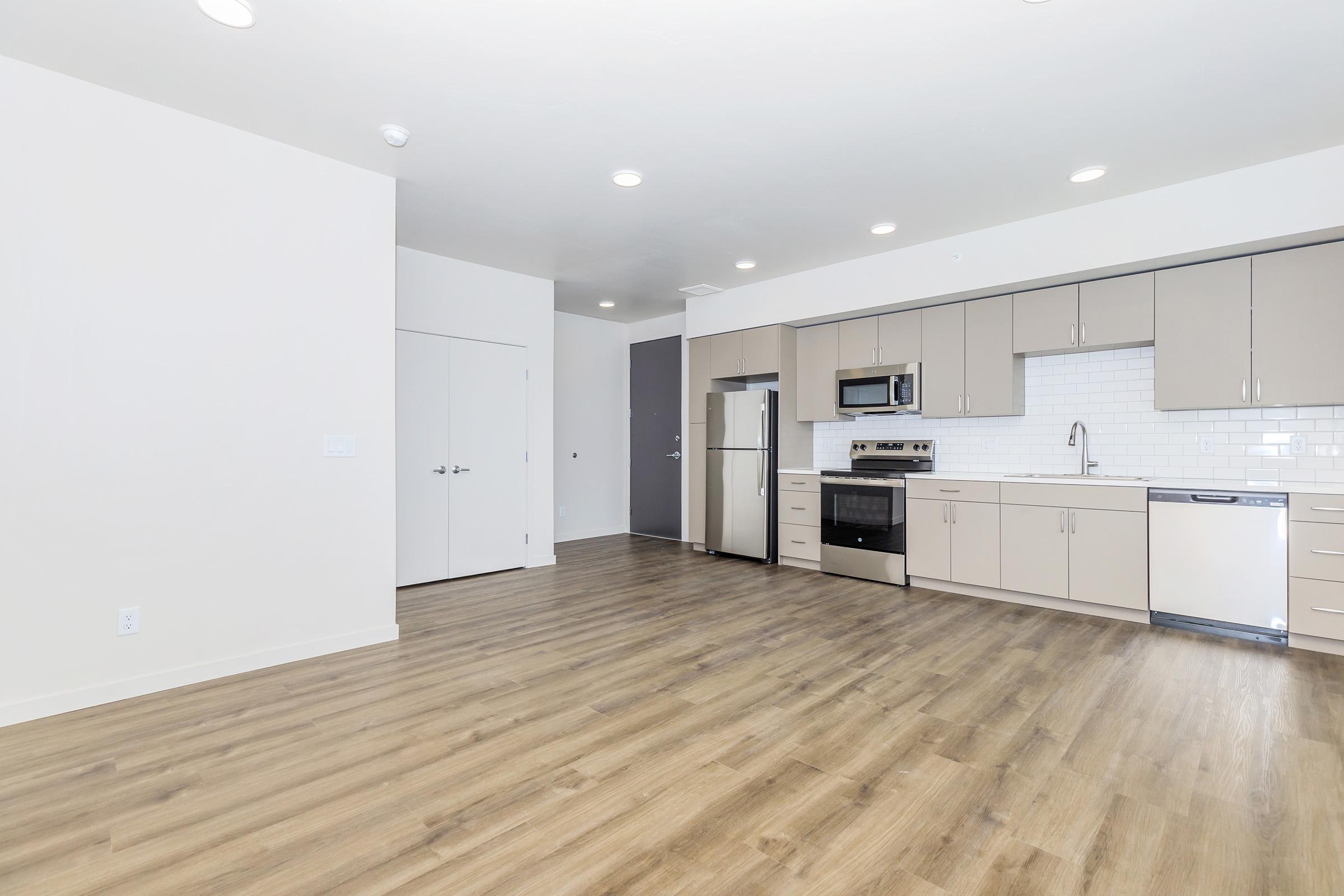 Modern kitchen and living area featuring sleek cabinets, stainless steel appliances, and a spacious layout with wood-like flooring. The kitchen includes a refrigerator, stove, microwave, and dishwasher, with a white tiled backsplash. Brightly lit space with white walls and a door leading to another room.