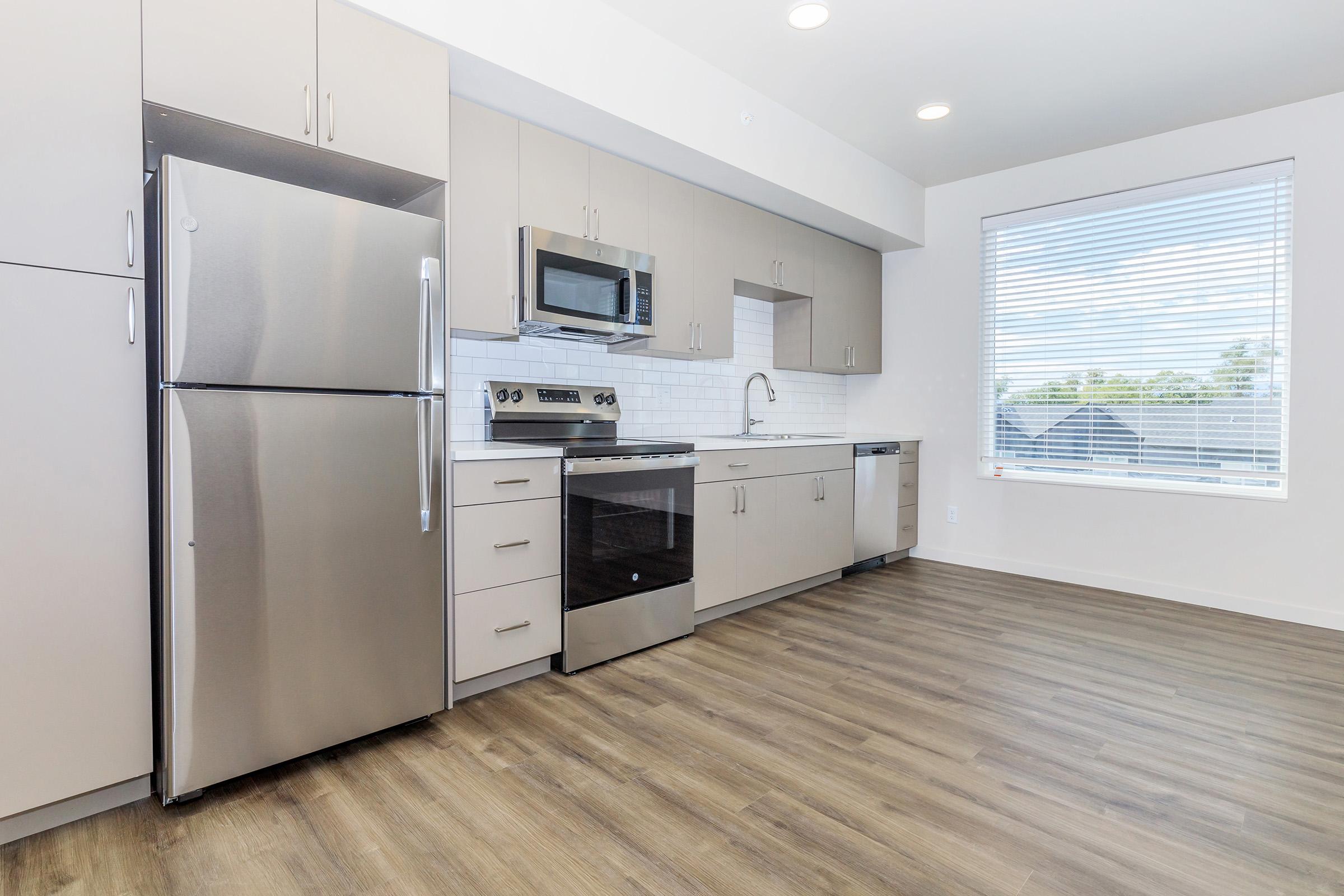 A modern kitchen featuring stainless steel appliances, including a refrigerator, microwave, and oven. The light-colored cabinets complement the sleek design. A large window with blinds illuminates the space, highlighting the hardwood floor and clean lines of the cabinetry.