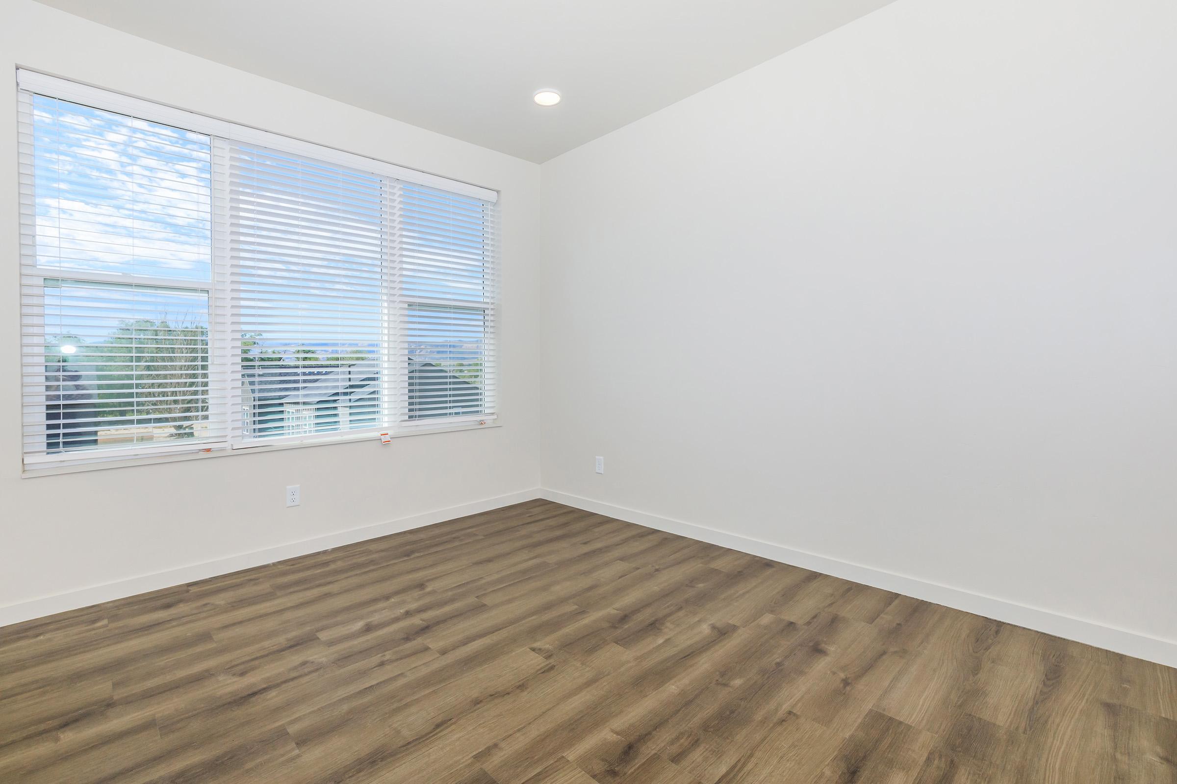 A spacious, empty room featuring light-colored walls and a large window with white blinds. The floor is made of wood laminate, providing a warm tone. Natural light filters through the window, illuminating the space, which is ready for decoration or use.