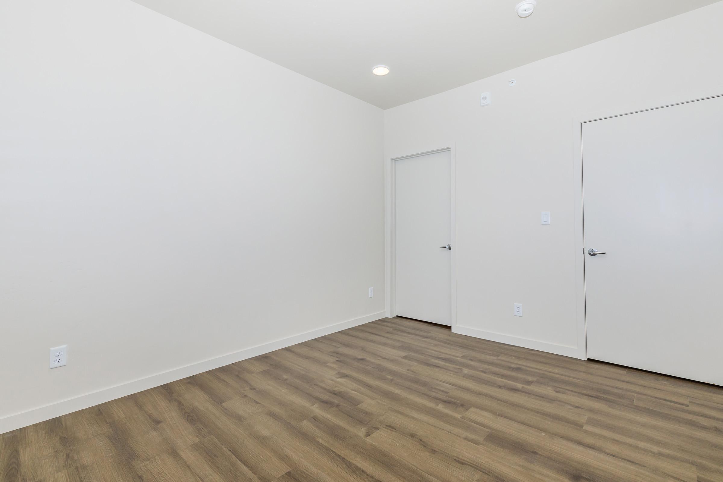 A minimalist, empty room with light-colored walls and a hardwood floor. There are two closed doors, one on the right side and one on the back wall. The space is well-lit with recessed ceiling lights, creating a bright and open atmosphere.