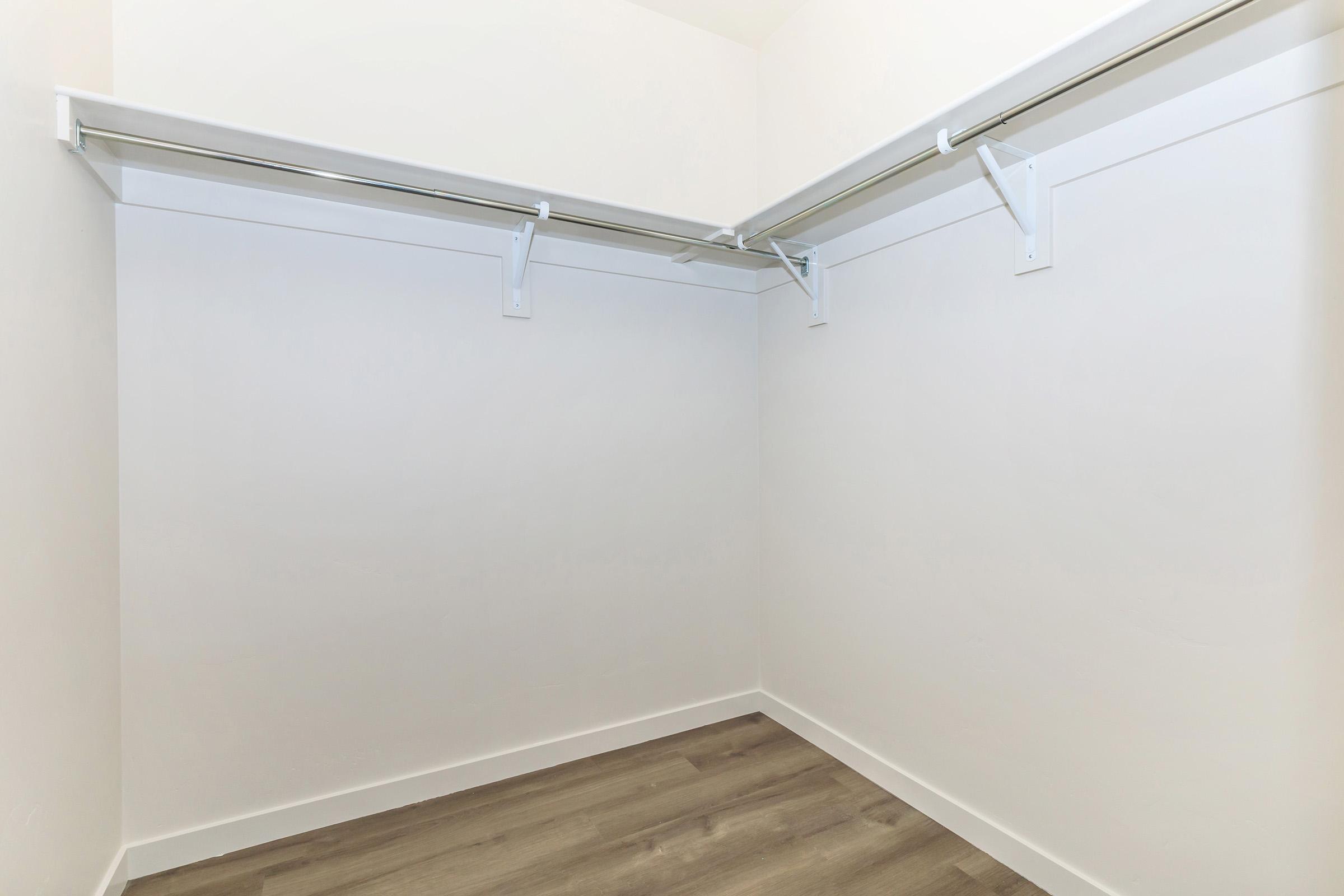 Empty closet space with white walls, a wooden floor, and two hanging rods. The corners of the room are visible, showcasing the open and uncluttered design, making it suitable for storage or organizing items.