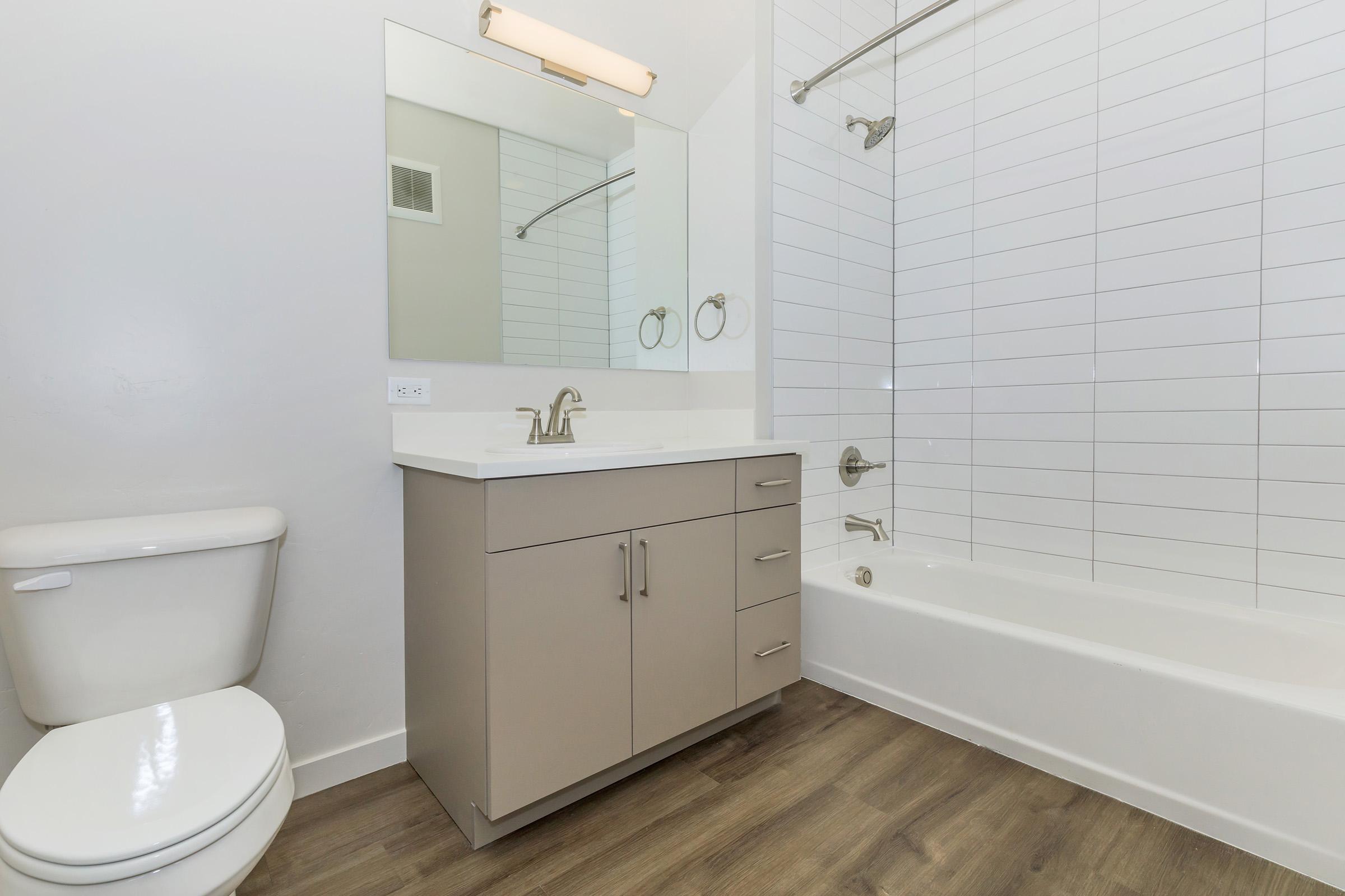 A modern bathroom featuring a white bathtub, a sleek vanity with drawers, a rectangular mirror, and a toilet. The walls are adorned with white tiles, and the floor is made of wood-like laminate. A shower fixture is present above the tub, creating a clean and inviting space.