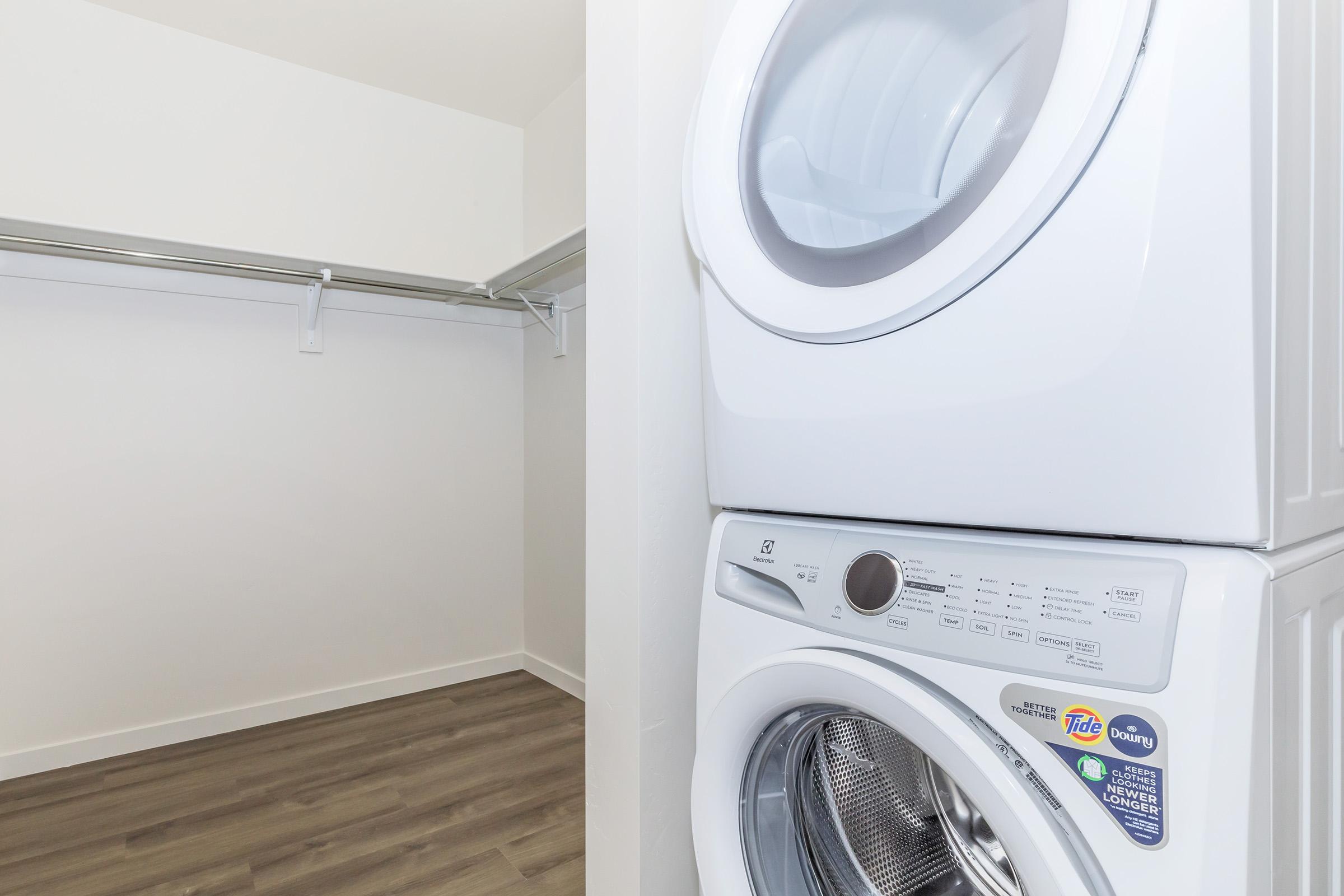 A modern laundry area featuring a stacked white washer and dryer unit. The background shows a clean, empty closet with a rod for hanging clothes. The floor is made of light wood-like material, creating a bright and spacious atmosphere.