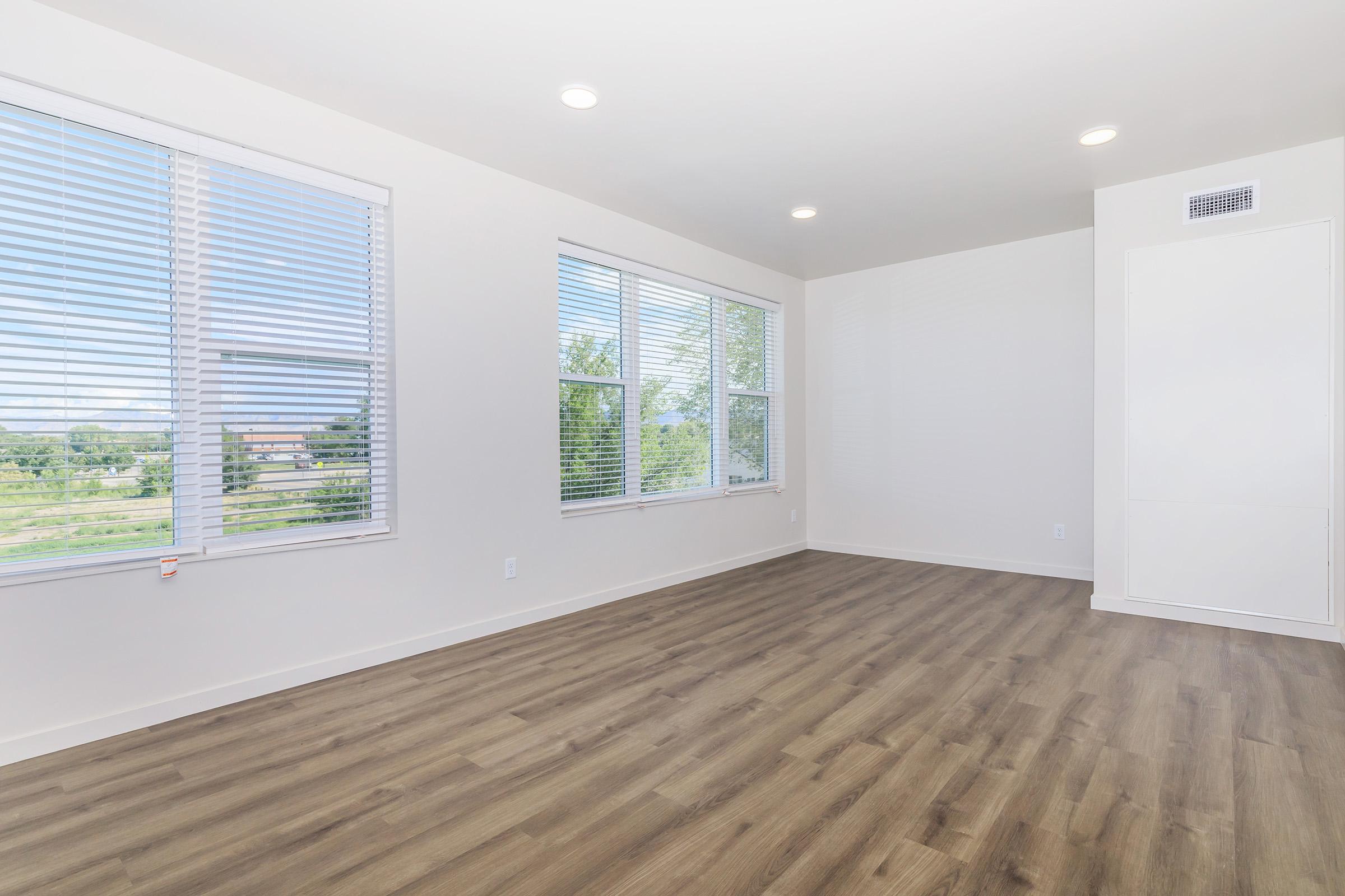 A bright, empty room featuring large windows with white blinds, light walls, and a smooth wooden floor. The space has recessed lighting and a clean, minimalistic design, with a view of greenery outside. There’s a built-in storage cabinet in the corner, creating an open and airy atmosphere.