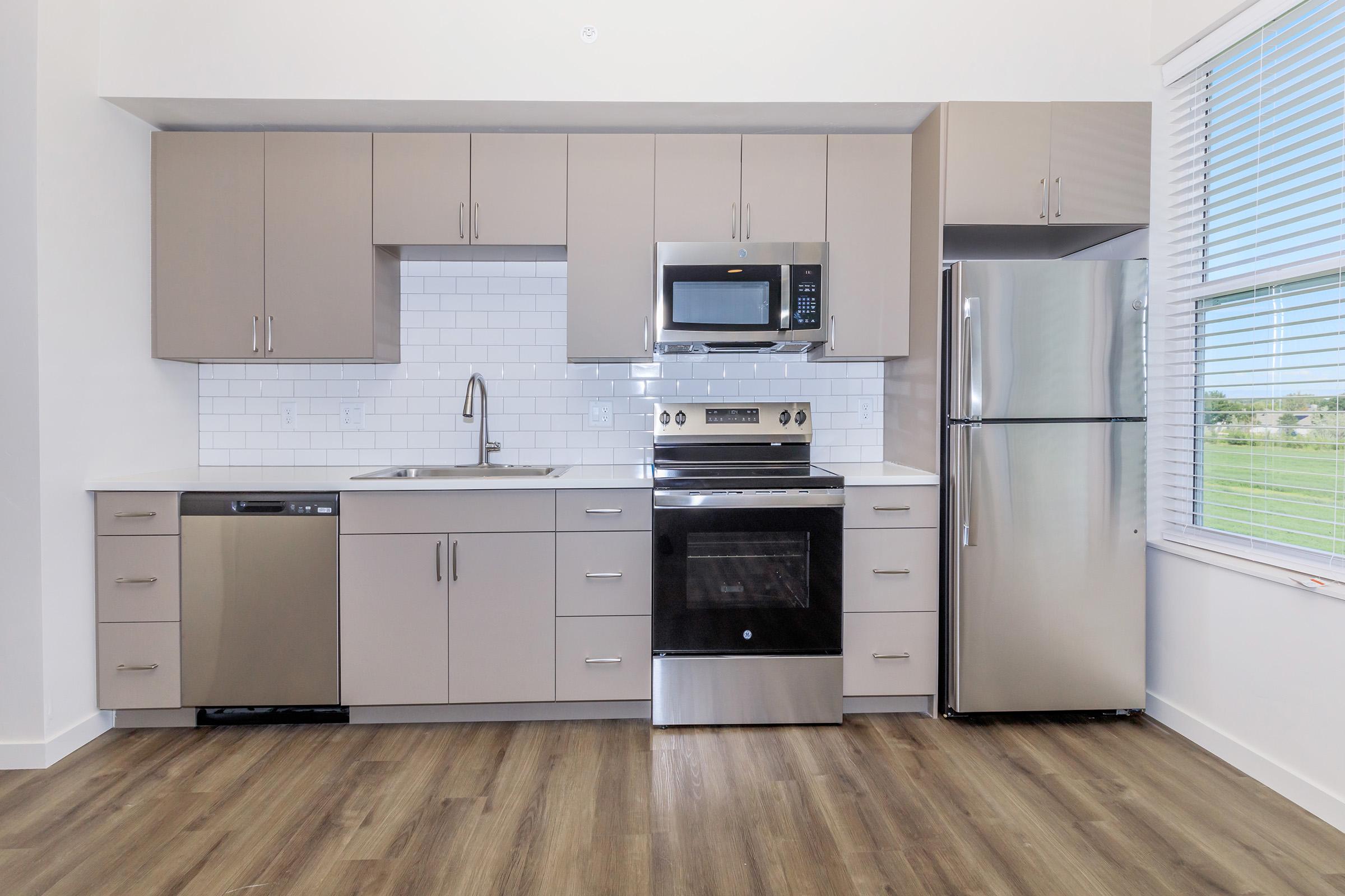 Modern kitchen featuring light gray cabinets, stainless steel appliances including a microwave, oven, refrigerator, and dishwasher. A white tile backsplash complements the countertops, while large windows provide natural light and a view of greenery outside. The floor is a warm-toned wood laminate.