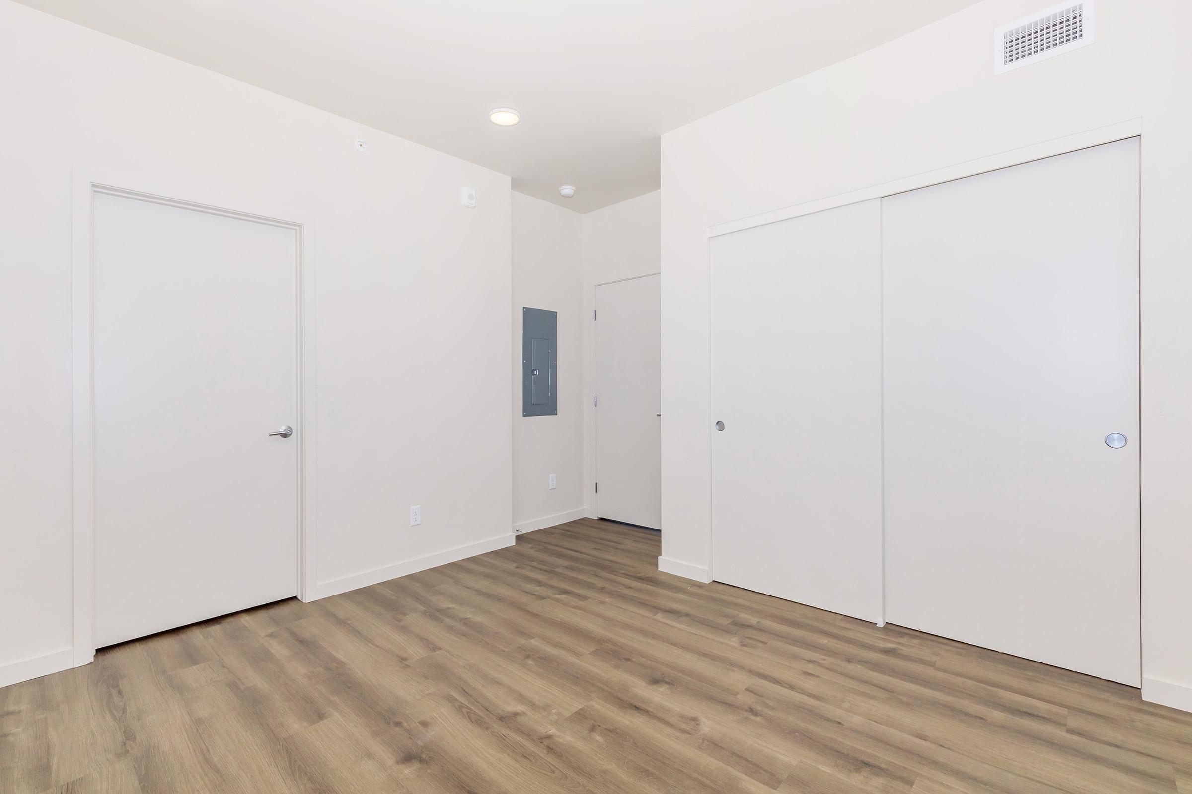 Empty room with light-colored walls and a tan wooden floor. There are two closed doors, one with a vertical electrical panel, and one leading to another space. The room has overhead lighting and no furniture, creating a spacious and minimalist appearance.