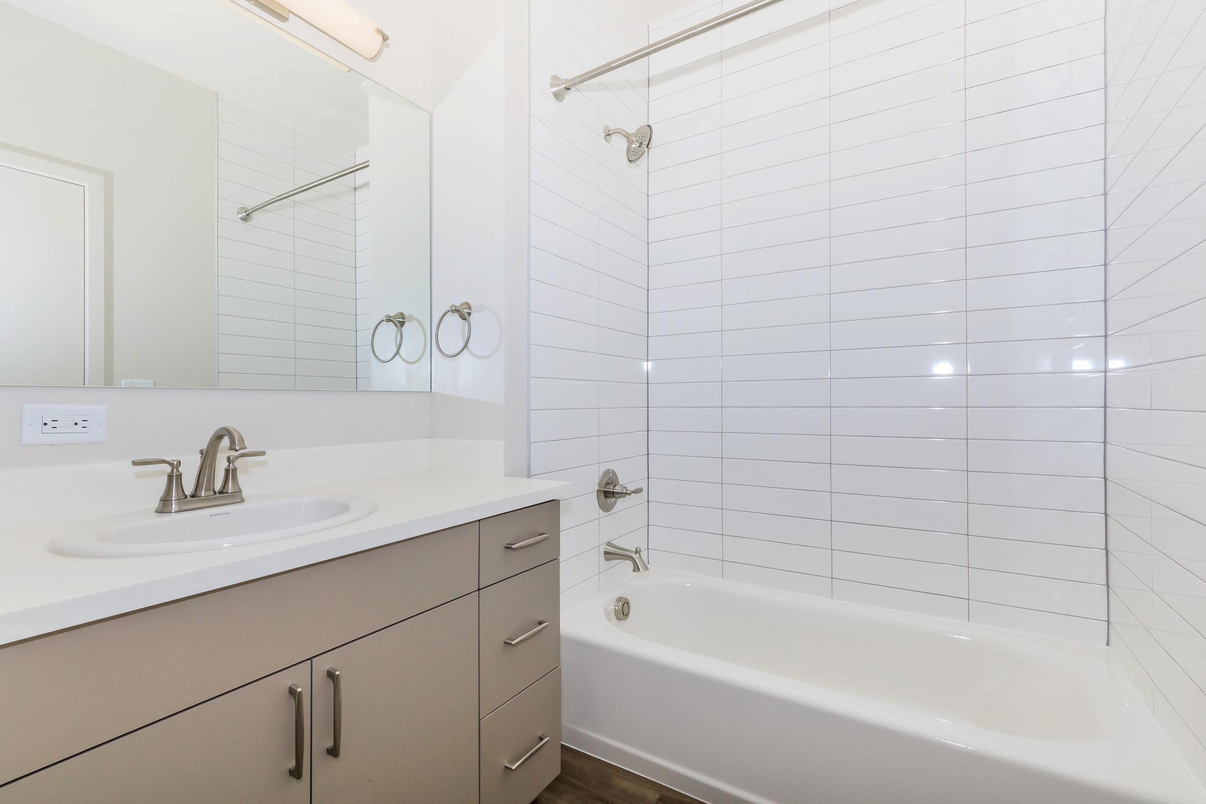 A modern bathroom featuring a clean design with a bathtub and shower combination. The walls are adorned with white rectangular tiles, and there is a sleek vanity with a circular sink and a mirror above it. Two towel rings are mounted on the wall, complementing the contemporary style.