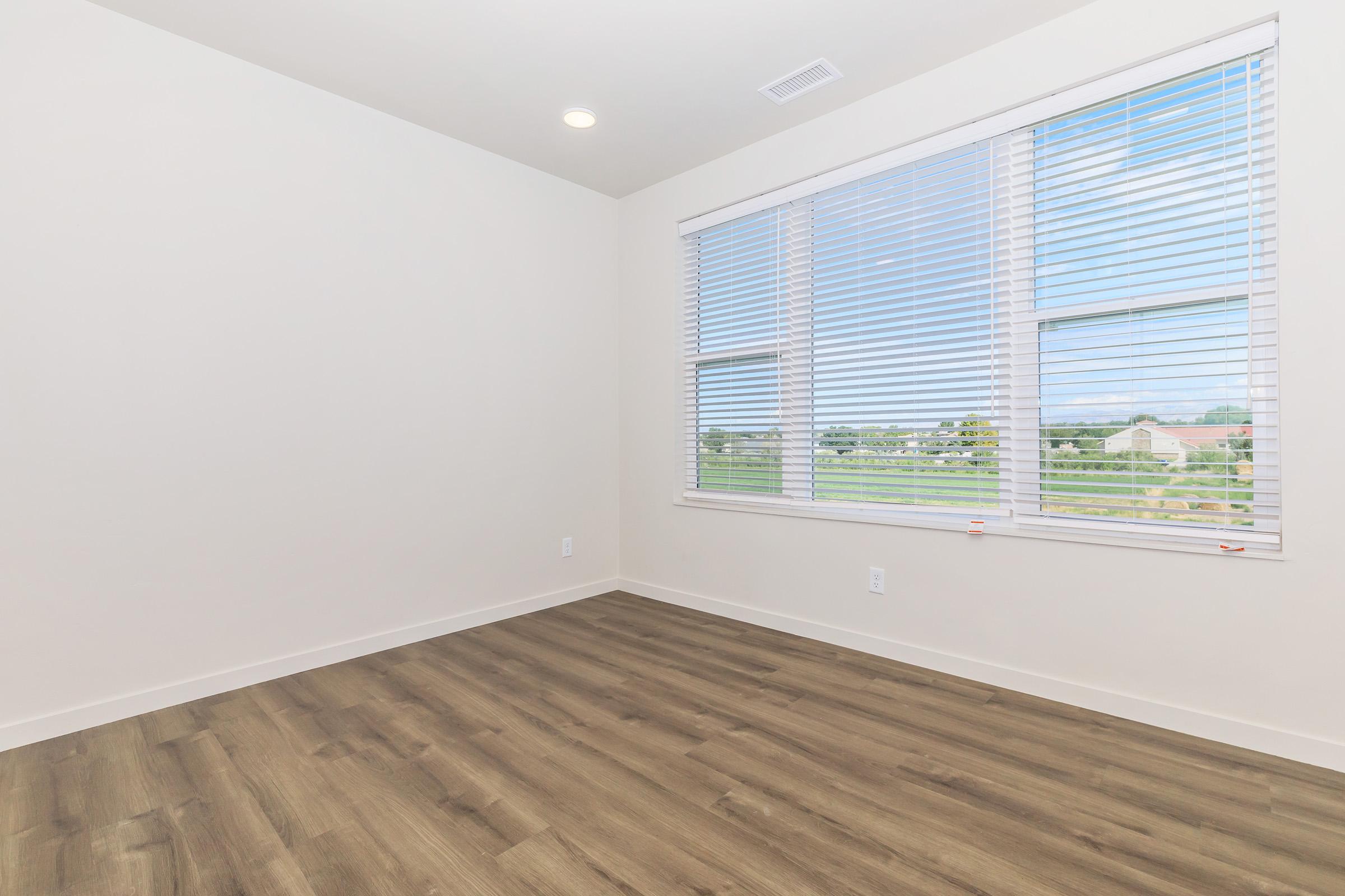 A clean, empty room with light-colored walls, large windows covered with blinds, and a wooden floor. The room has a spacious and bright appearance, with a view of an outdoor landscape visible through the windows.