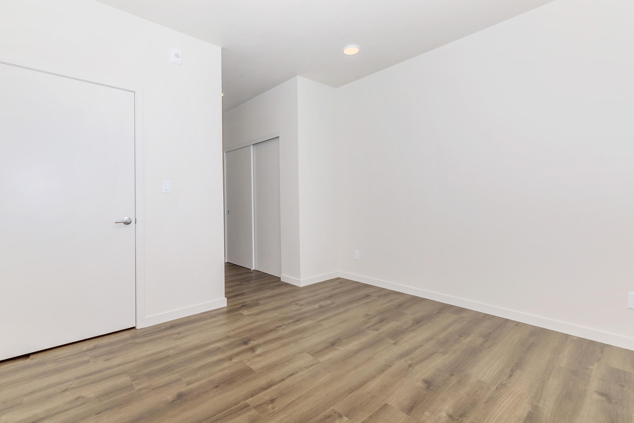 A bright, empty room with light-colored walls and a warm wooden floor. There is a closed white door on the left, and a sliding closet door is visible further along the wall. The space has a minimalistic design, with ample natural light entering the room from an out-of-view source.