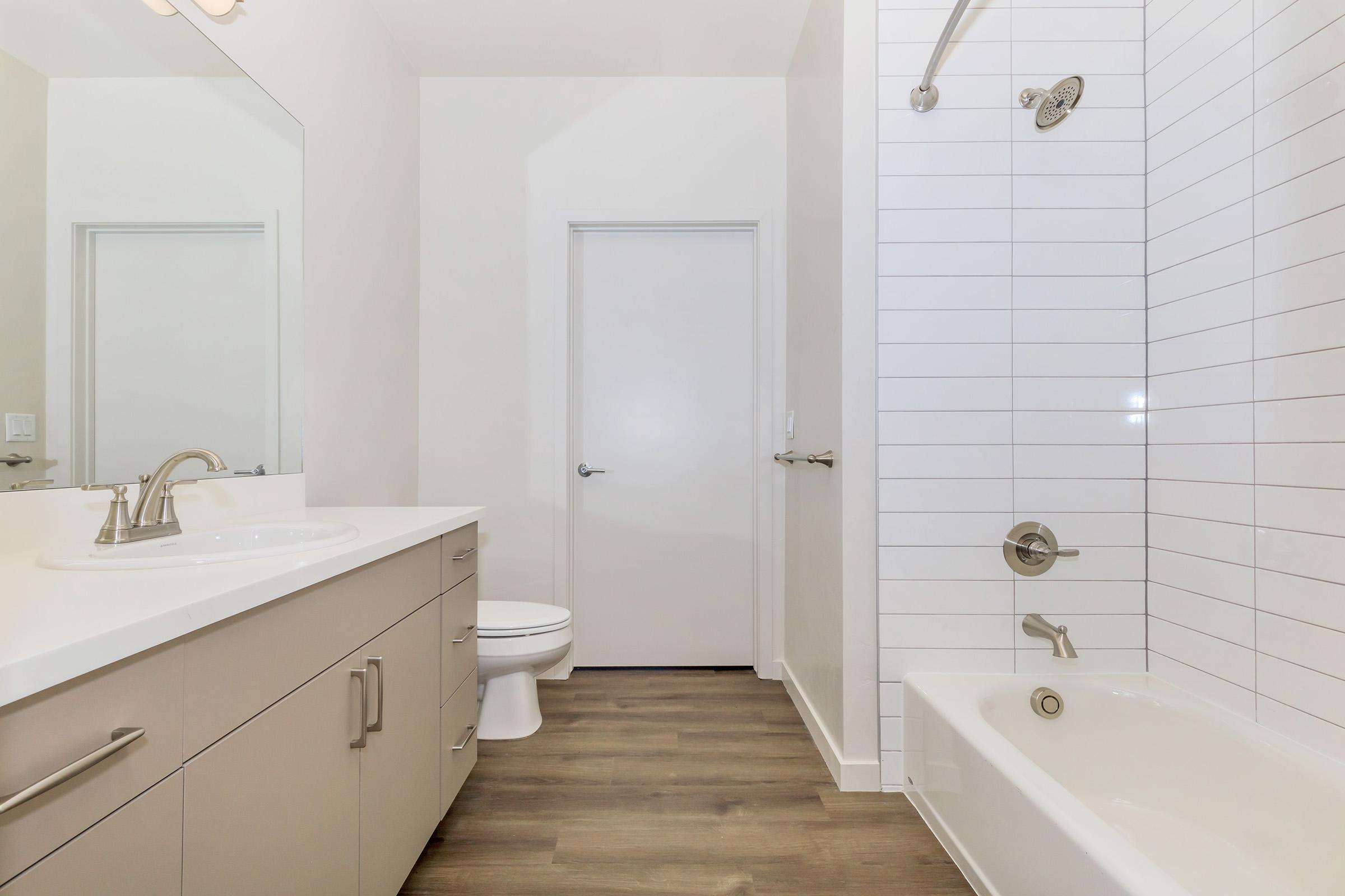 A modern bathroom featuring a double sink vanity with a large mirror, a white toilet, a shower tub combo with a sleek faucet, and a tiled wall. The floor is wooden, and there is a minimalist door leading to another room. The overall design is clean and contemporary.