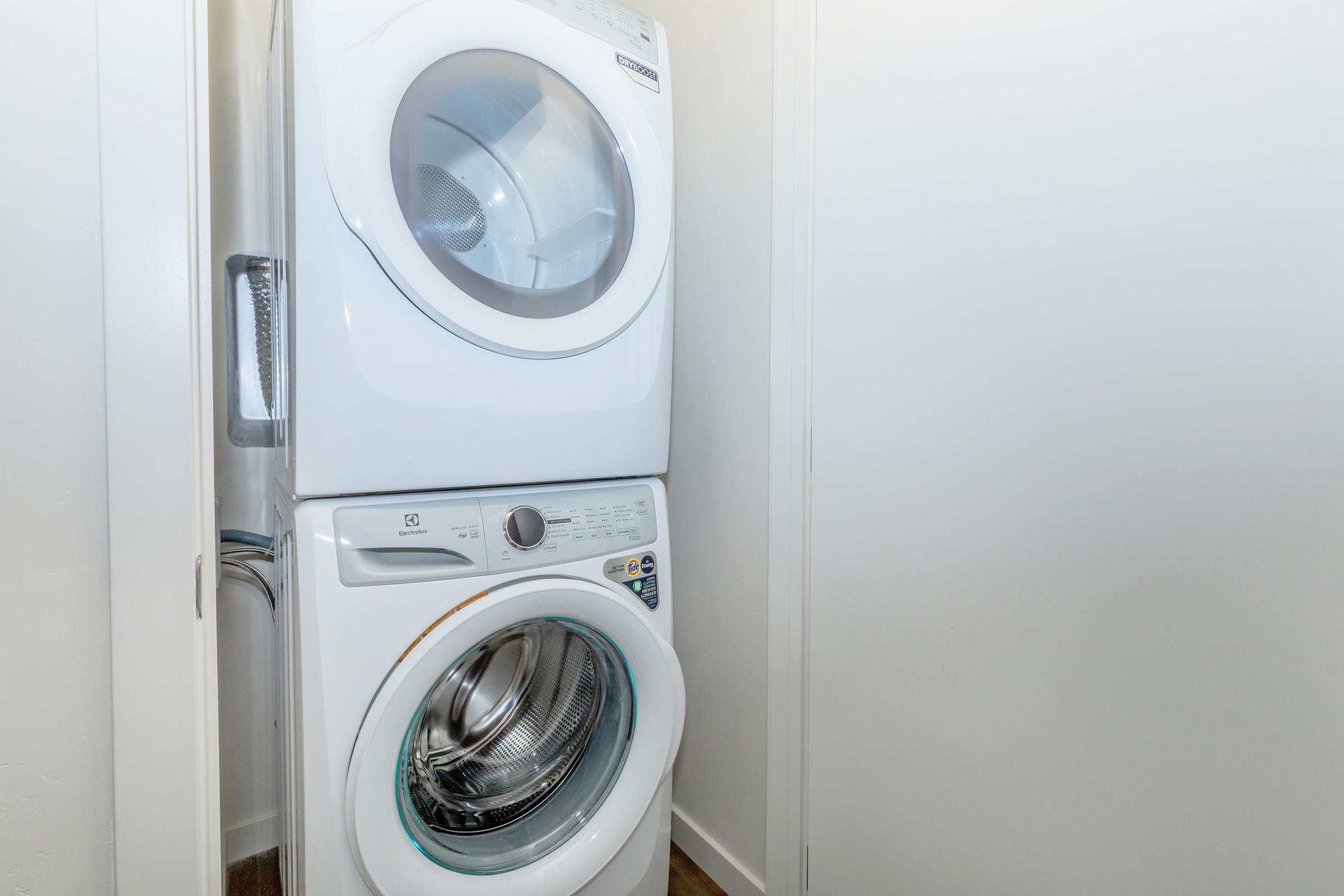 A stacked washer and dryer unit located in a narrow laundry space with a light-colored wall. The washer is on the bottom and the dryer on top, both featuring modern designs and controls. The floor is laminate, and there is minimal additional decor in the area.