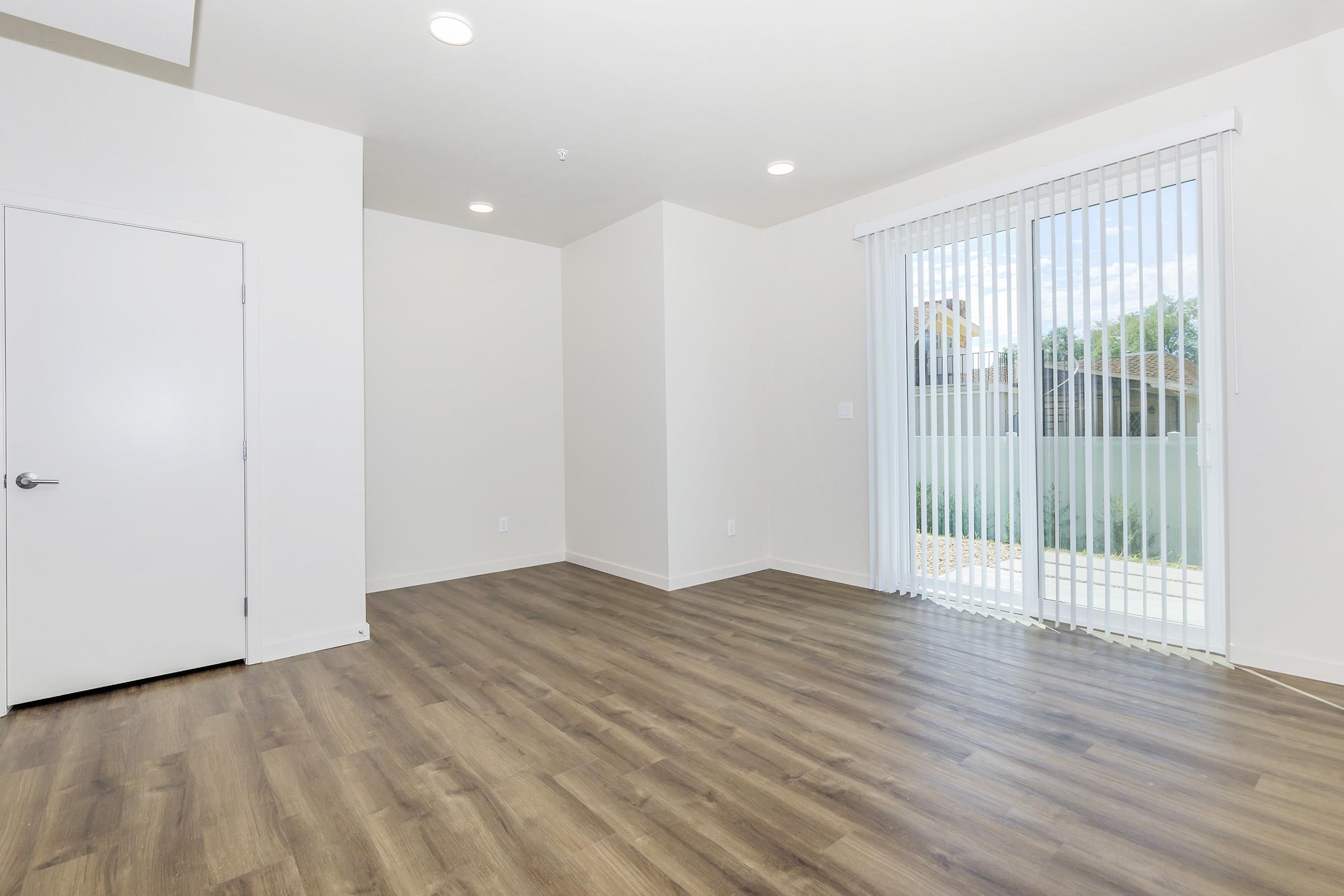 A bright, empty room with light-colored walls and a wooden floor. On the right, there are vertical blinds covering a large window that opens to an outdoor area. A single door is on the left side, and the overall space has a clean and modern feel.