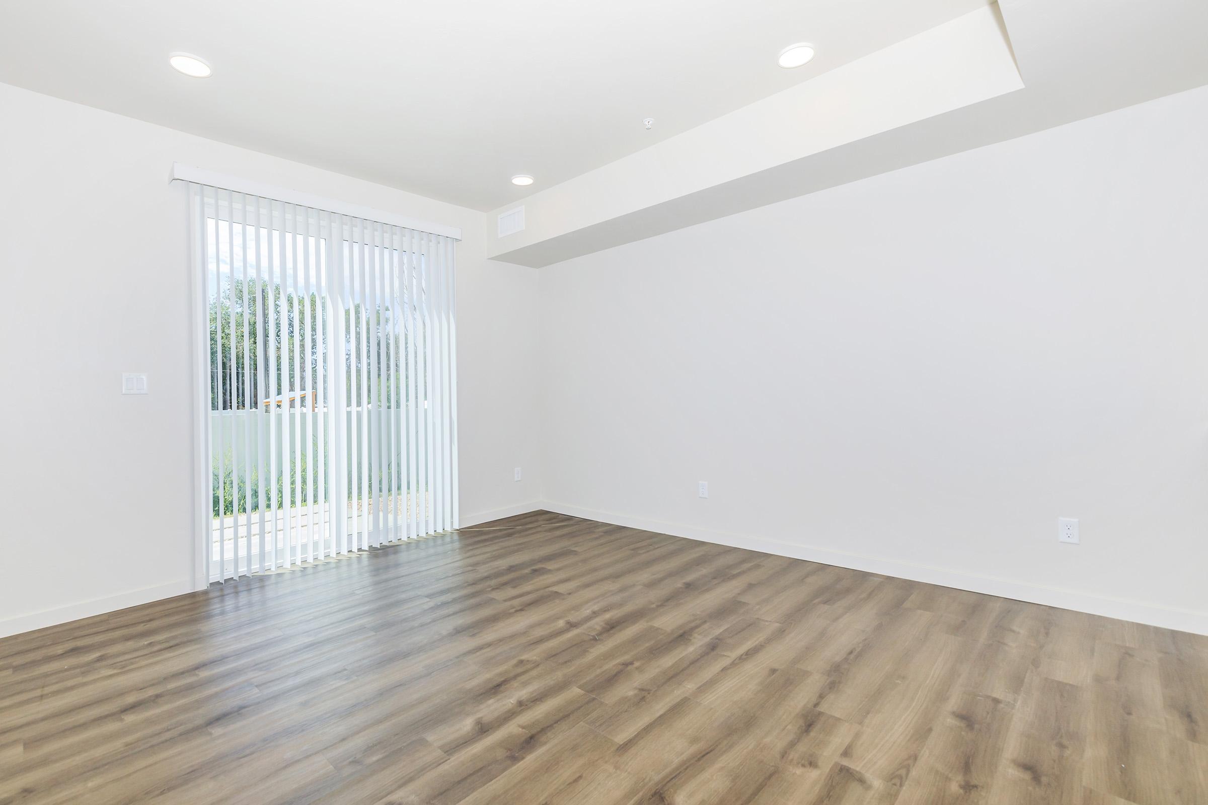 A bright, empty room with light-colored walls and wood-style flooring. There is a large sliding glass door with vertical blinds leading to an outdoor area, allowing natural light to fill the space. The ceiling is simple with recessed lighting, giving a modern feel. No furniture is present.