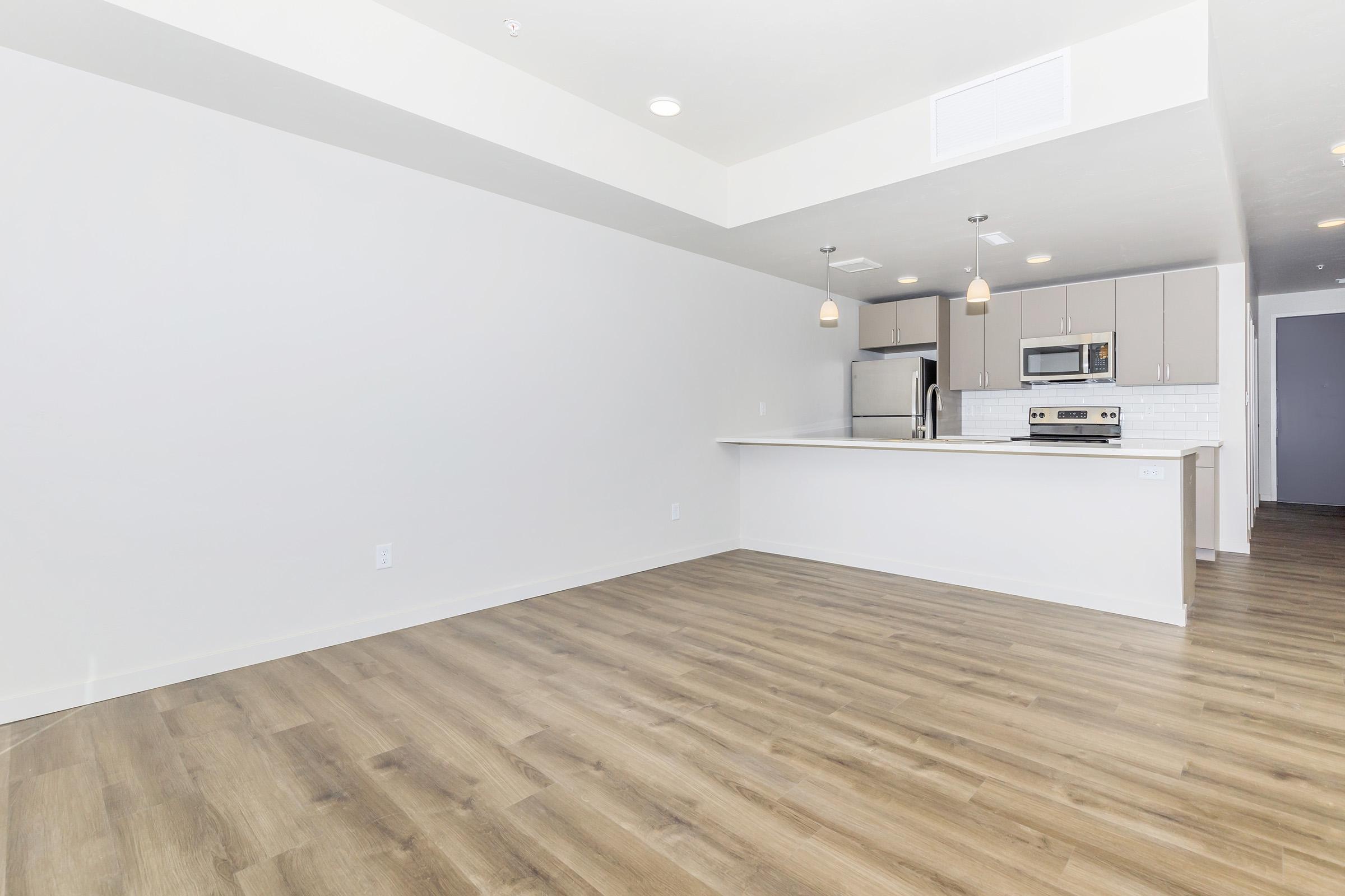 A spacious and modern interior of an empty apartment, featuring light wood flooring, a kitchen with stainless steel appliances, a white countertop, and minimalist cabinetry. The walls are painted light gray, and the ceiling is bright with fixture lights. A doorway leads to another room in the background.