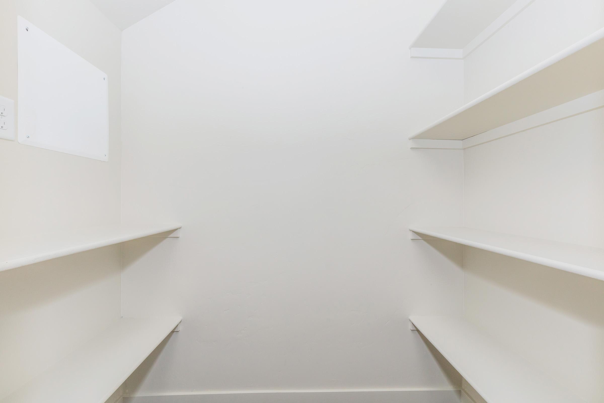 An empty pantry with bare white shelves on either side, positioned in a corner. The walls are a light neutral color, and there is a blank white square on one wall, possibly for a light switch or decoration. The overall space appears clean and minimalistic.