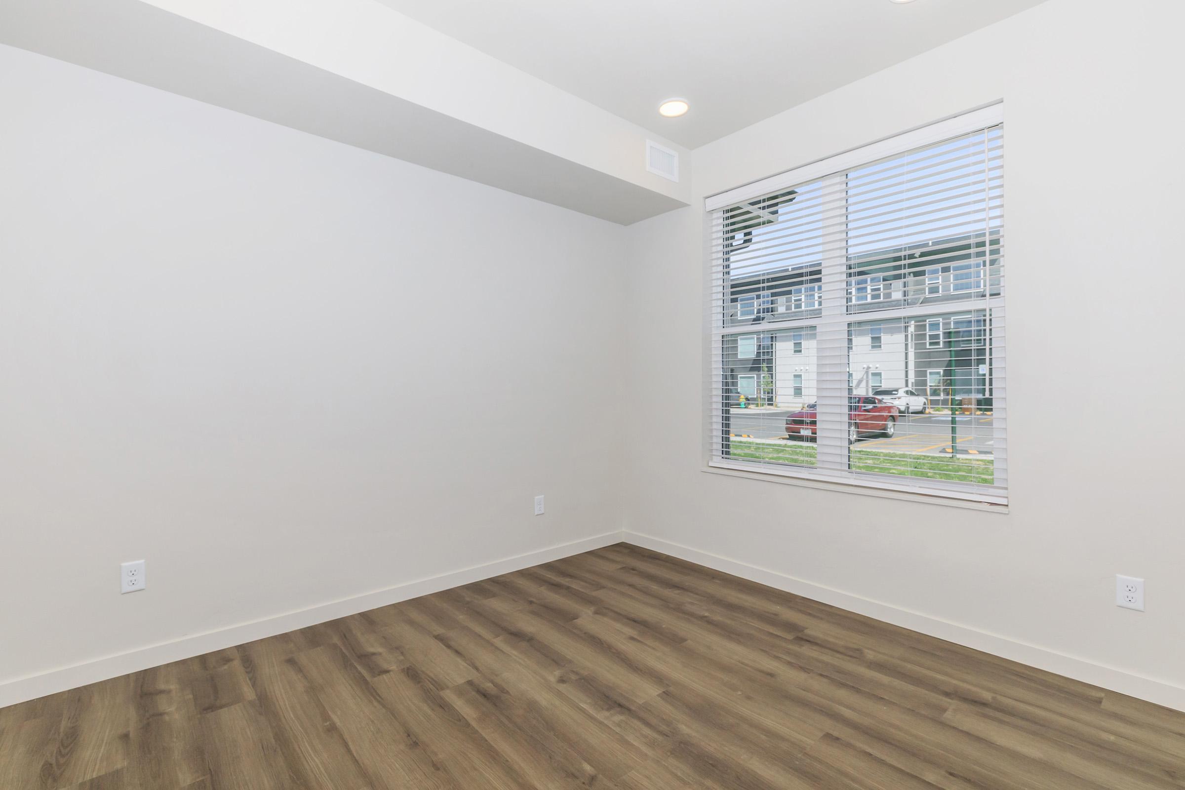 A brightly lit empty room with light-colored walls and wooden flooring. There is a large window with white blinds that overlooks an outdoor area with vehicles parked outside. The room has no furniture and appears spacious and modern.