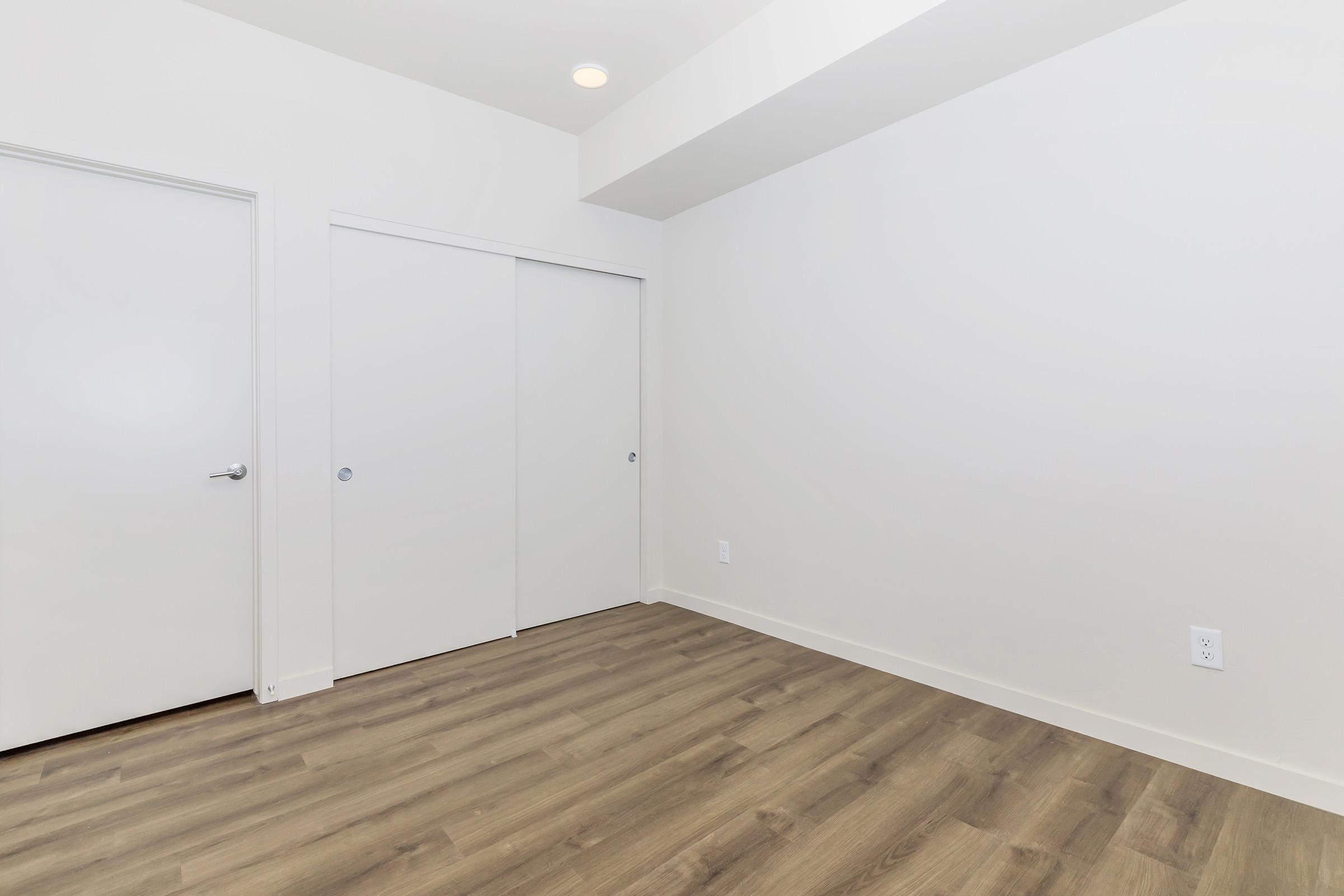 An empty room with light-colored walls and a wooden floor. There are two closed doors on the left side, one with a handle and both blending into the wall. The ceiling has a recessed light, and the space is well-lit and uncluttered, creating a minimalist and modern aesthetic.