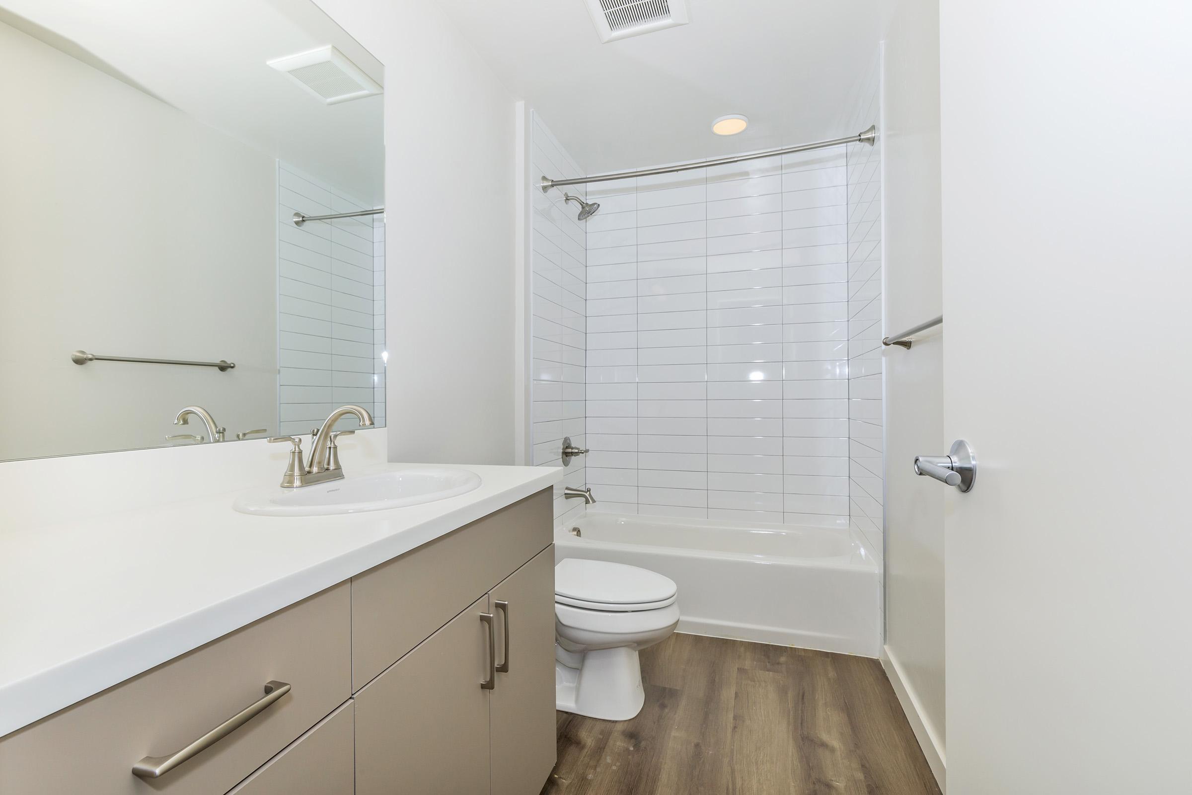 A clean and modern bathroom featuring a bathtub with a shower, a white sink with a sleek faucet, and beige cabinetry. The walls are tiled in light colors, and the floor has a wood-like finish. Bright lighting enhances the spacious feel of the room.