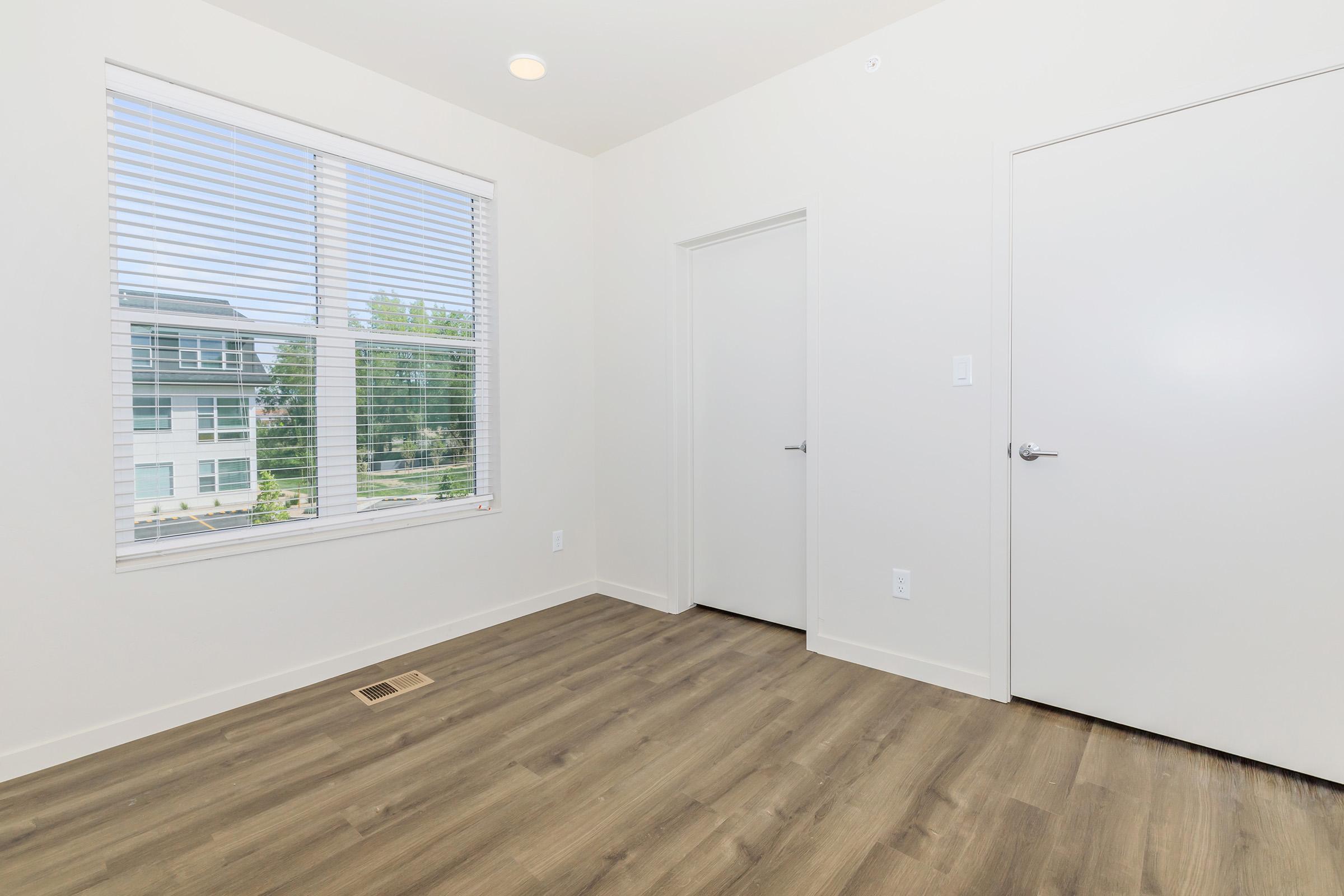 A well-lit, empty room with a large window featuring horizontal blinds. Light-colored walls and a modern door are visible. The floor is covered with light wood laminate. The room has a clean and minimalist design, creating a spacious feel.