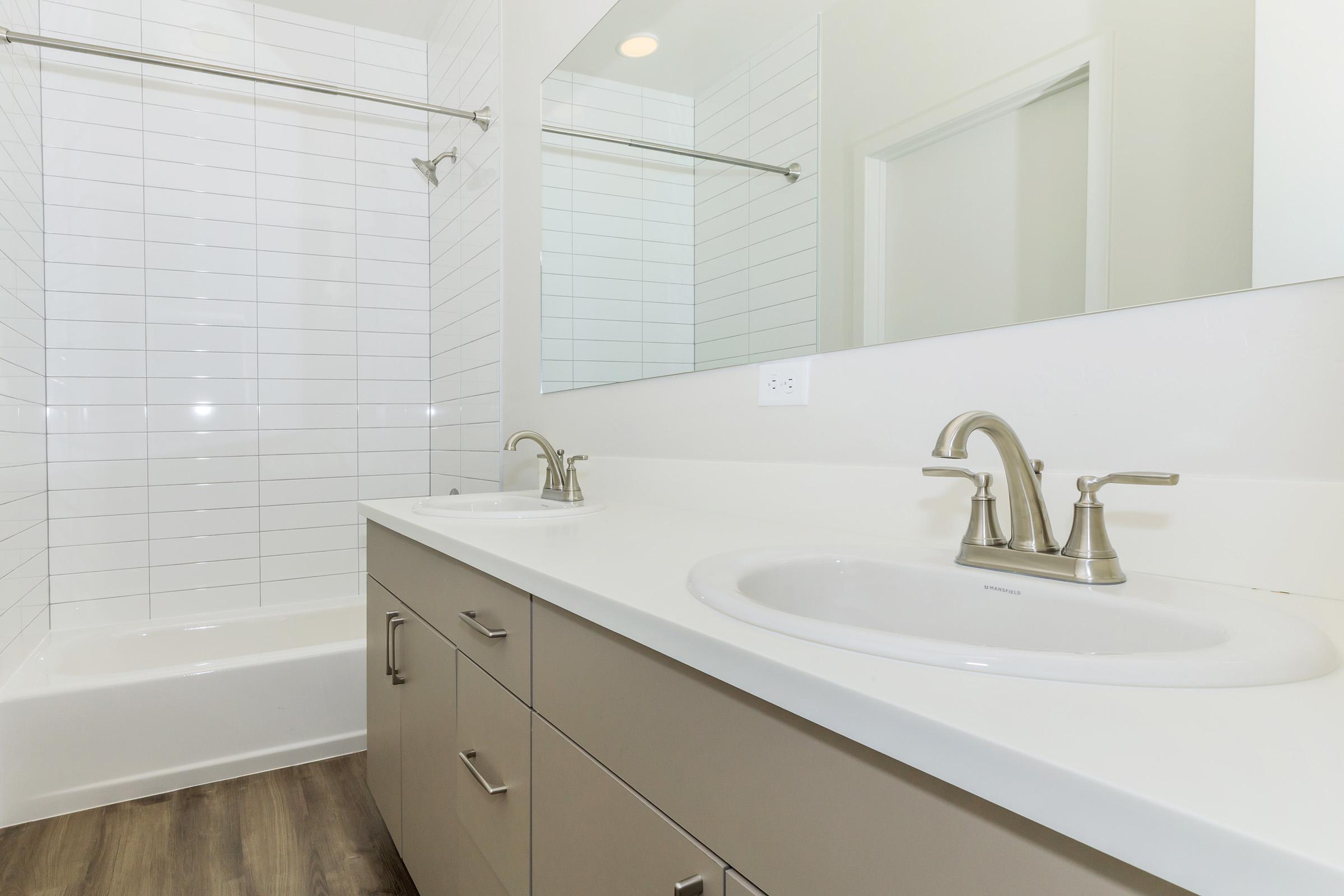 A modern bathroom featuring a double sink vanity with brushed nickel faucets, a large mirror above, and a white tiled wall. There's a bathtub with a shower fixture and a light-colored cabinet underneath the sinks. The flooring is a warm-toned wood look, enhancing the clean and spacious design.