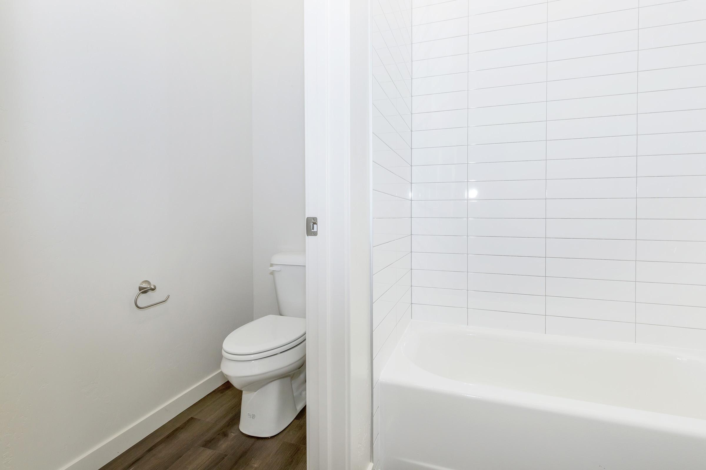 A clean, minimalist bathroom featuring a white toilet and a bathtub with a tiled wall. The floor is wooden, and there is a towel bar mounted on the wall next to the toilet. The overall design is simple and modern, with neutral colors and plenty of natural light.