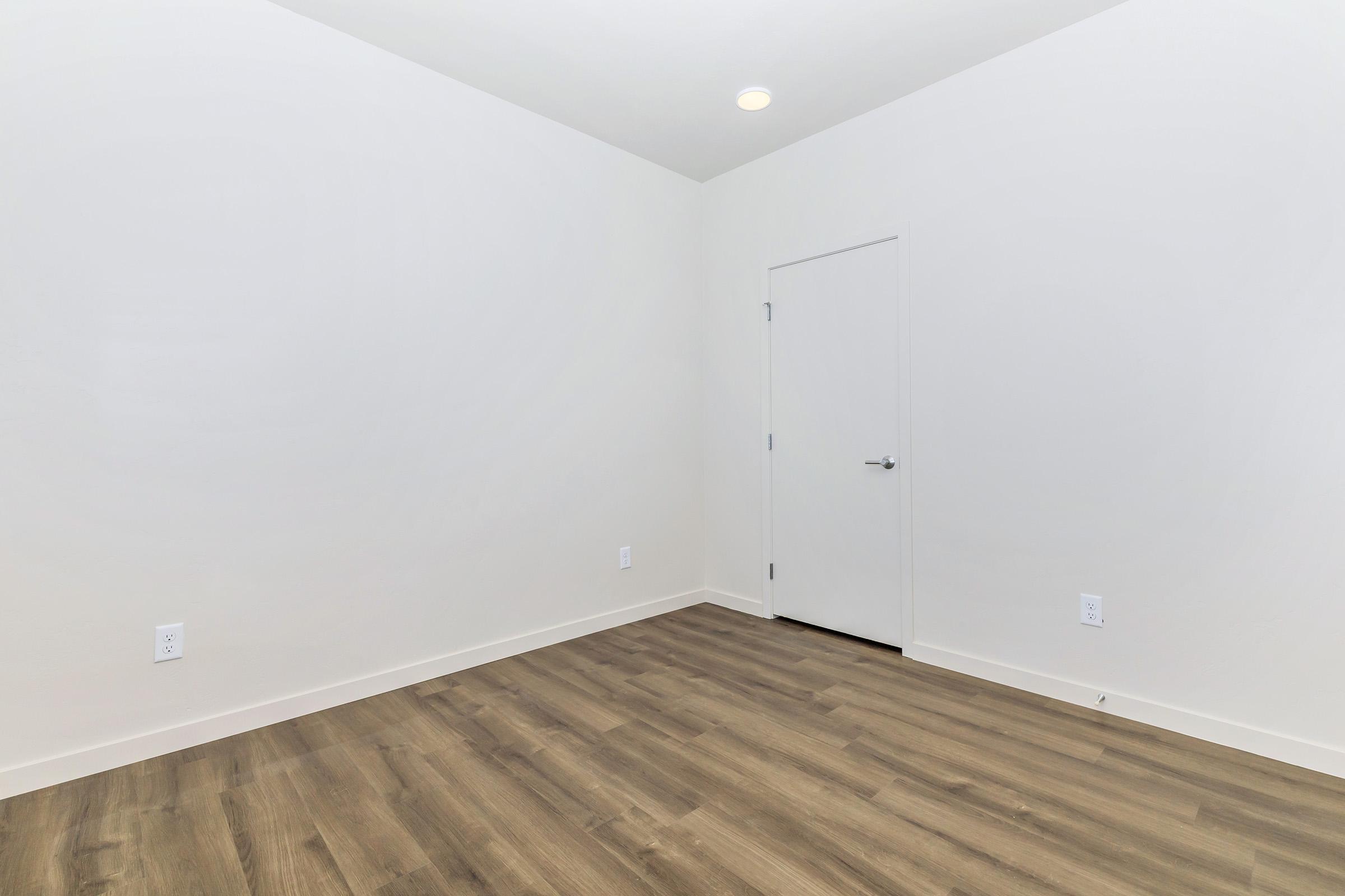 Empty room with light-colored walls and a wooden floor. There is a white door on one side, and the space is well-lit with a ceiling light. No furniture or decorations are present, giving a minimalistic appearance.