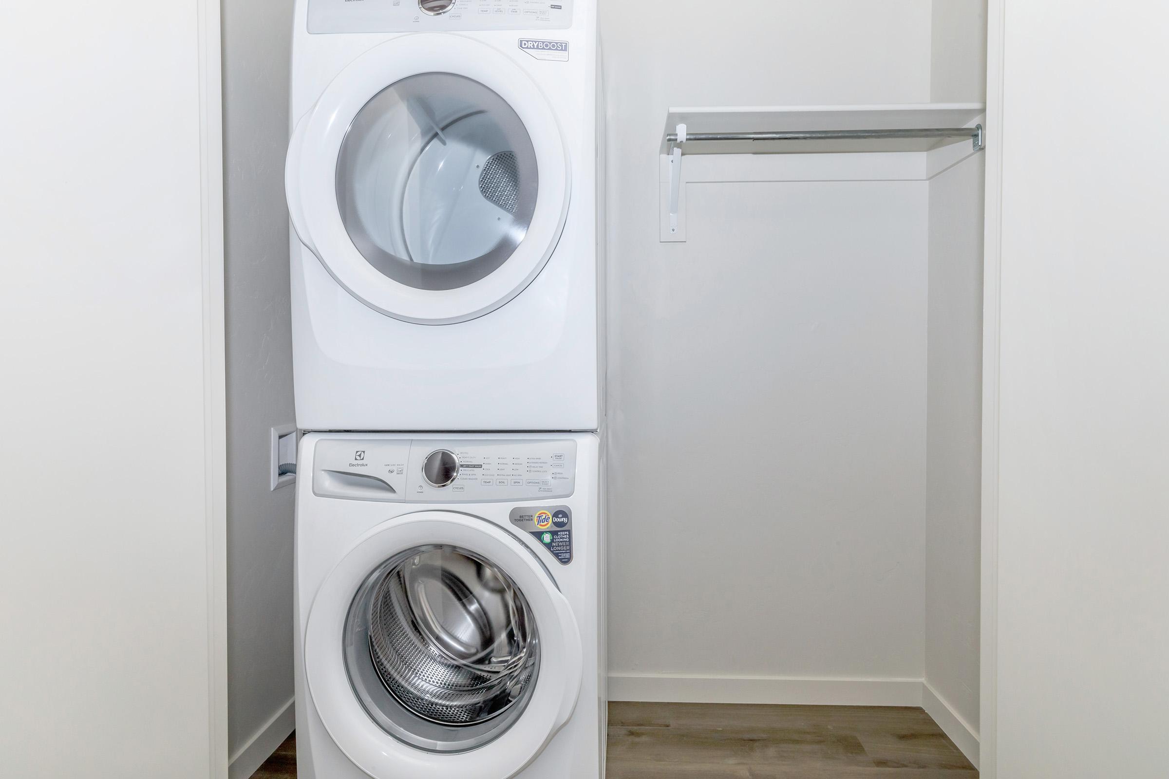 A stacked washer and dryer set in a bright, minimalistic laundry space. The appliances are white and located in a compact area with a simple wall shelf above them. The floor is light-colored, enhancing the clean and tidy look of the room.