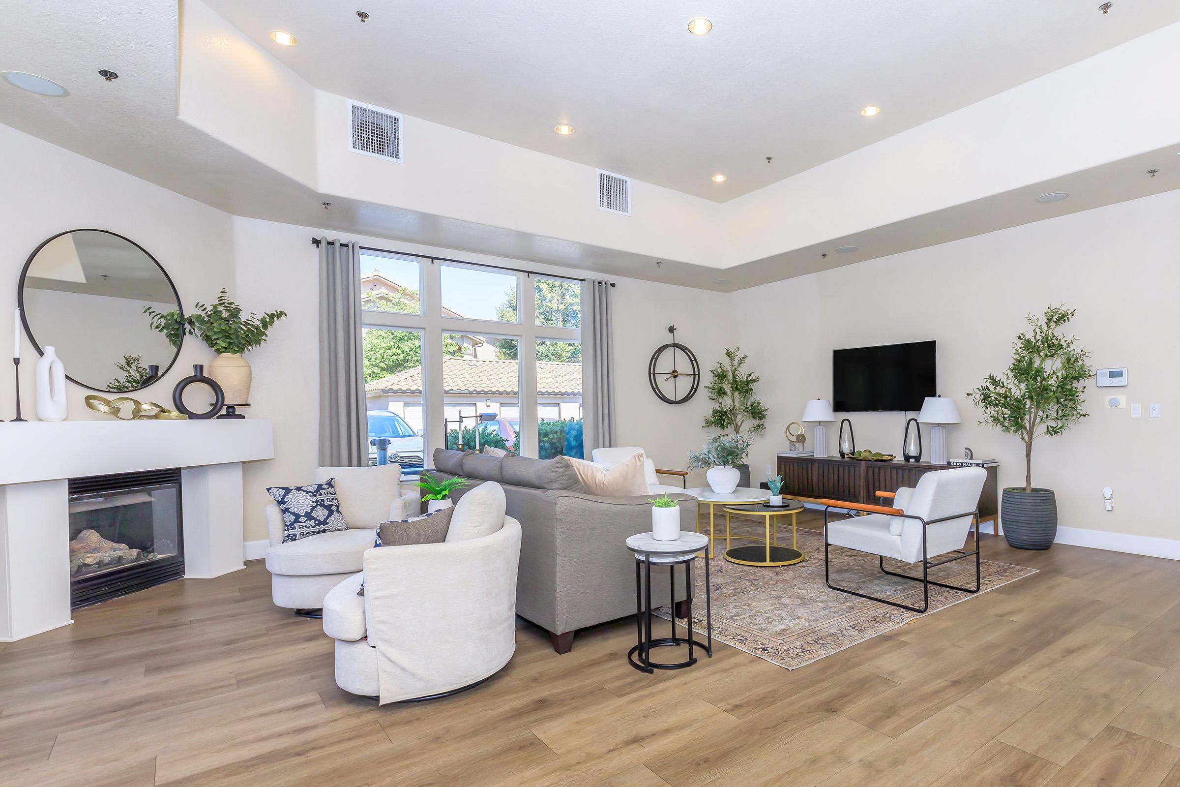 A modern living room featuring a cozy seating area with a gray sofa, armchairs, and a round coffee table. There’s a minimalist decor with plants, a fireplace, and a large window allowing natural light. A television is mounted on the wall, and a neutral color palette adds to the inviting atmosphere.