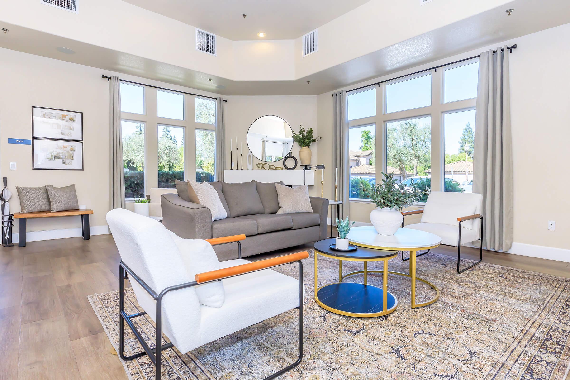A modern living room featuring a light-colored sofa, two white armchairs with wooden accents, and a round coffee table. Large windows allow natural light, while decorative plants and a mirror enhance the inviting atmosphere. A patterned area rug adds warmth to the space.