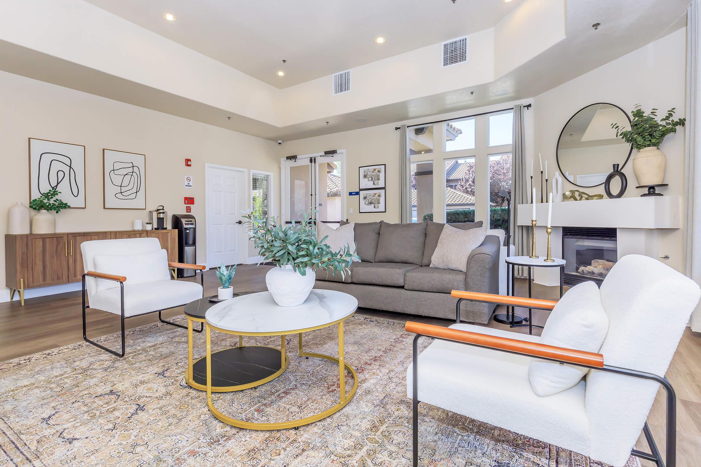 A stylish living room featuring a modern gray sofa, white armchairs, and a round coffee table with a potted plant. The space includes decorative wall art, a large mirror, and a cozy atmosphere with natural light streaming in through windows. A patterned rug adds warmth to the decor.