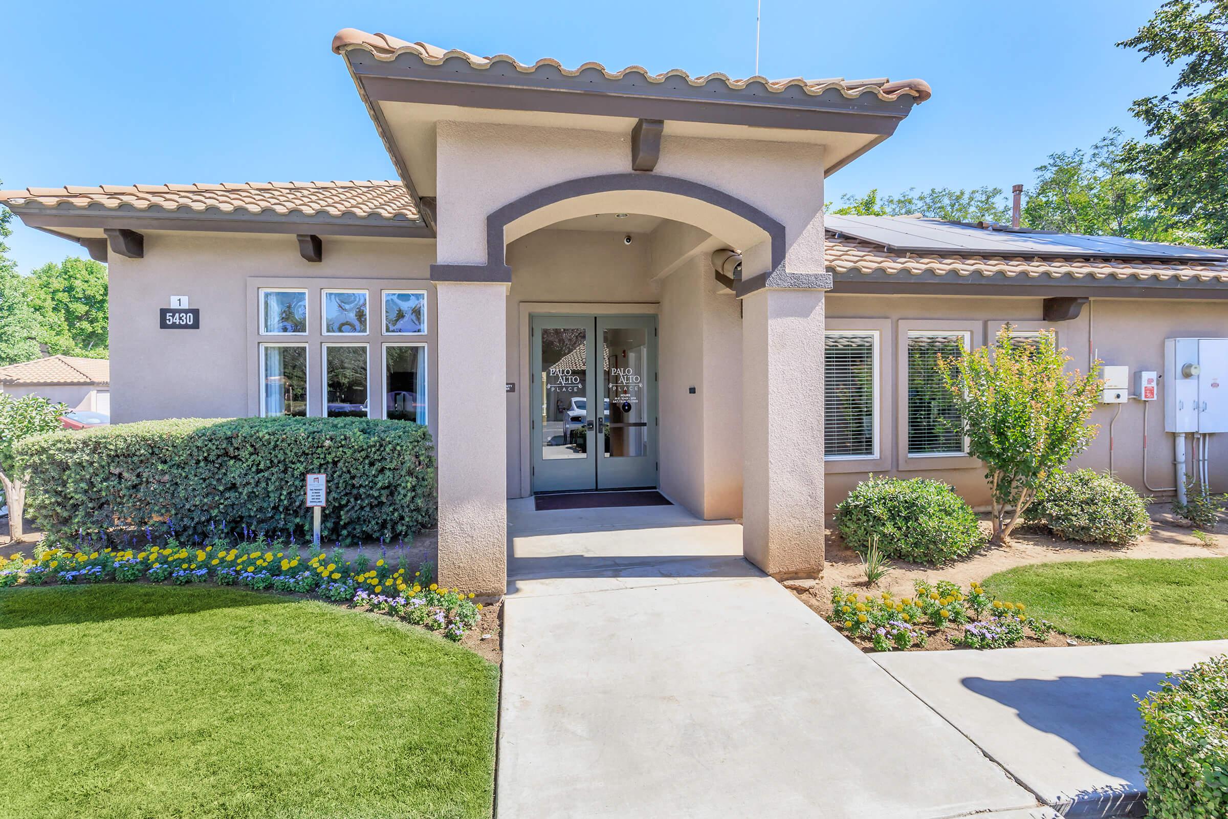 A beige-colored building with a tiled roof and large front windows. The entrance features double glass doors framed by an archway. A well-maintained walkway leads up to the door, lined with vibrant flower beds and manicured grass. Green shrubs and trees surround the exterior, contributing to a welcoming atmosphere.