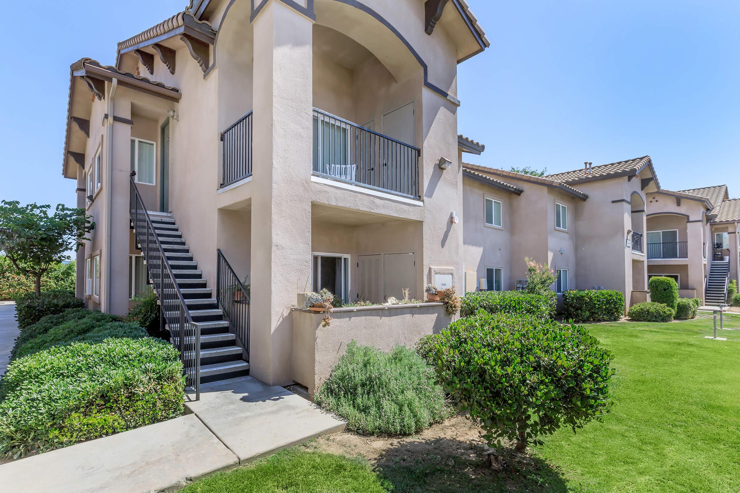Two-story residential building with beige stucco exterior and balconies. A set of stairs leads to the second floor. Surrounding greenery includes well-manicured lawns and shrubs. Clear blue sky in the background highlights the architecture and landscaping.