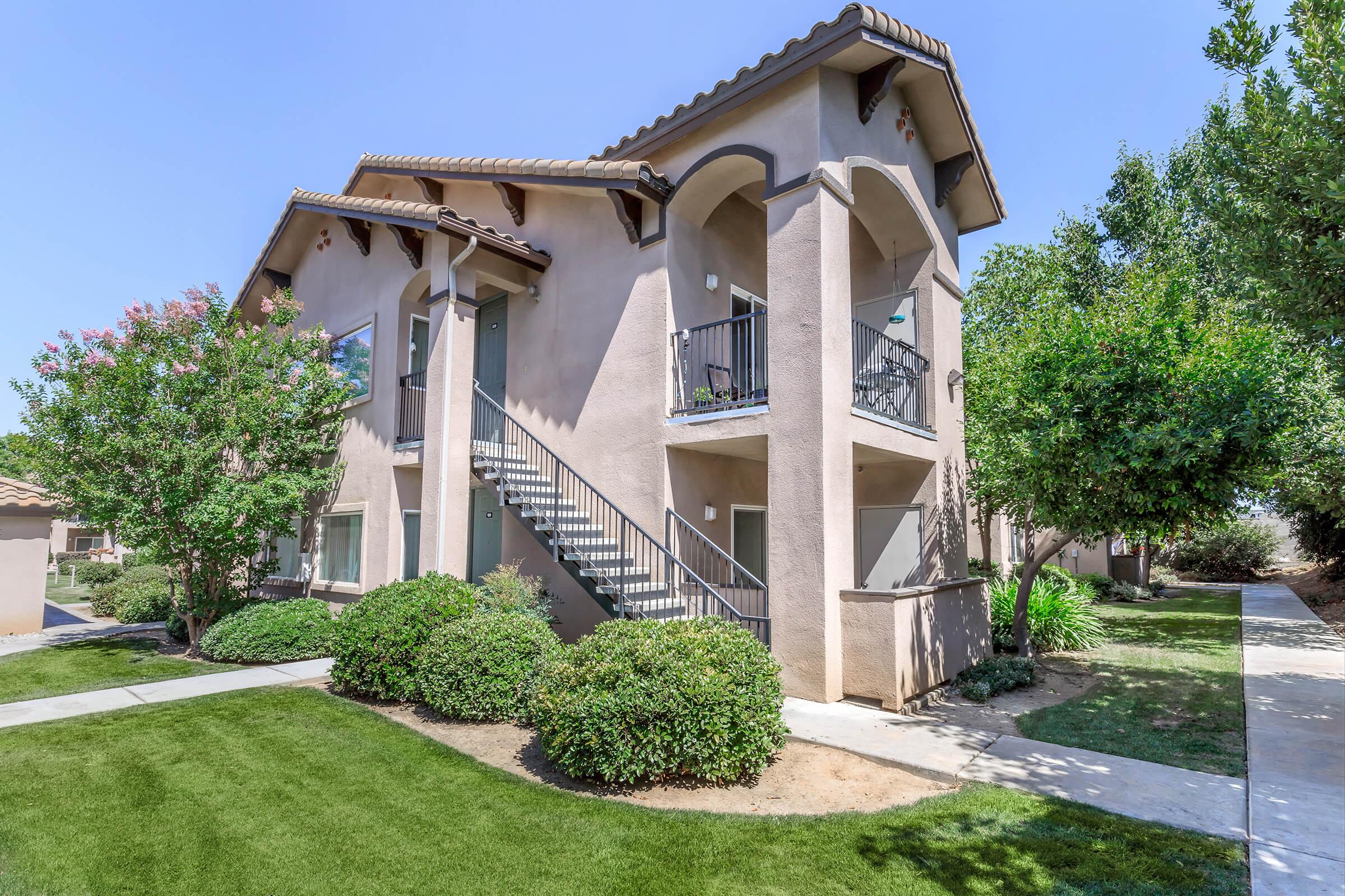 Two-story residential building with beige exterior. It features a staircase on the side, green shrubs, and trees surrounding the property. A well-maintained lawn and paved walkway are visible in front, with clear blue skies above. The overall scene conveys a serene and inviting atmosphere.