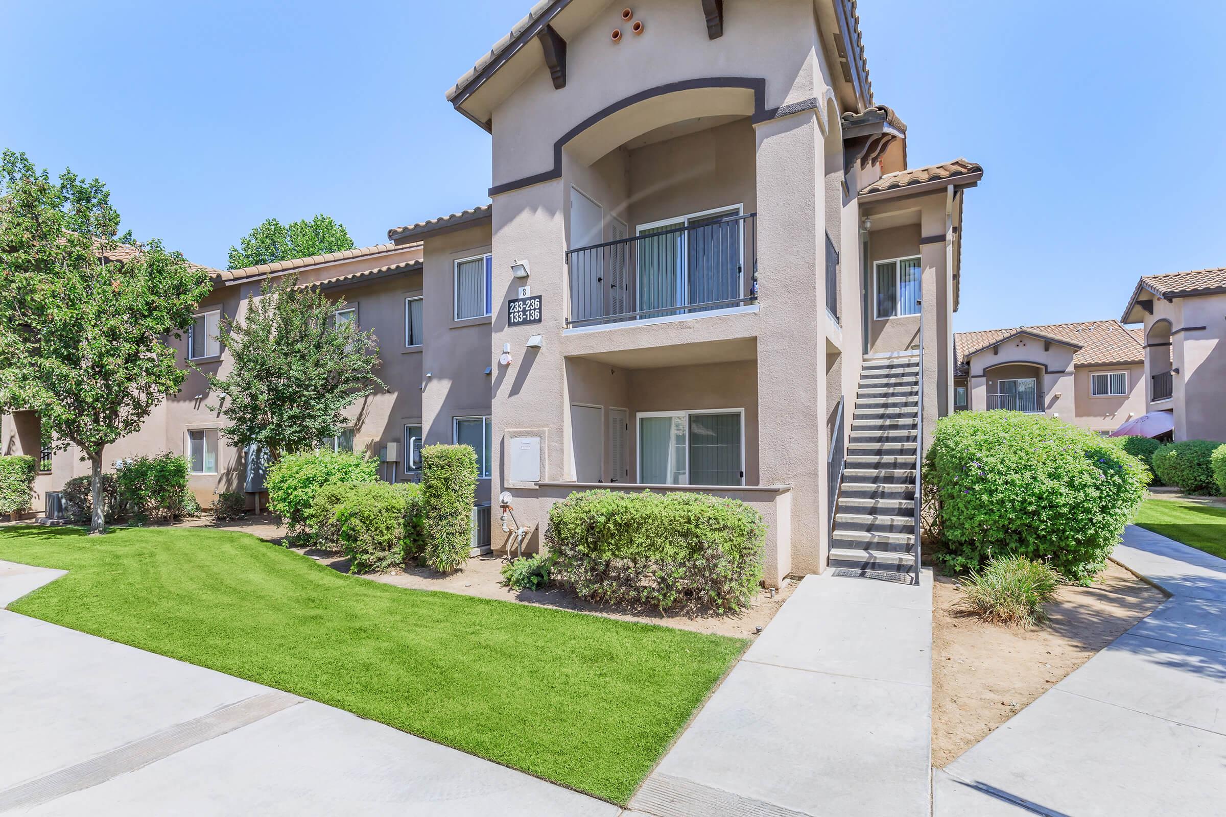 Exterior view of a multi-unit residential building featuring a beige facade, a staircase leading to an upper-level apartment, lush green landscaping, and well-maintained pathways. Bright blue sky in the background, giving a sunny and inviting atmosphere.