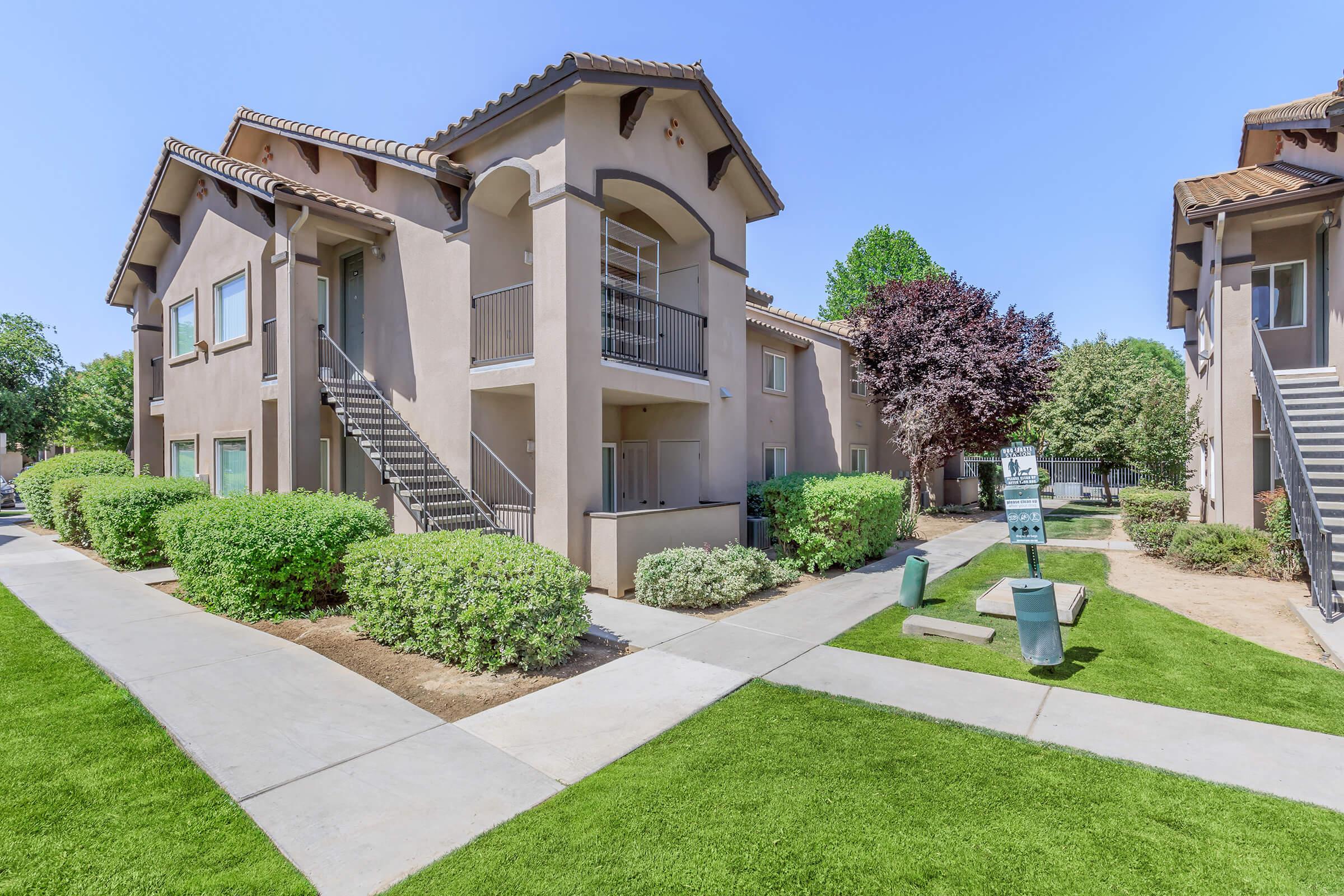 Two-story residential apartment building with a light brown exterior, featuring balconies and a stairway. Lush green bushes and trees surround the property, with a manicured lawn and a paved pathway leading to the entrance. Bright blue sky overhead.